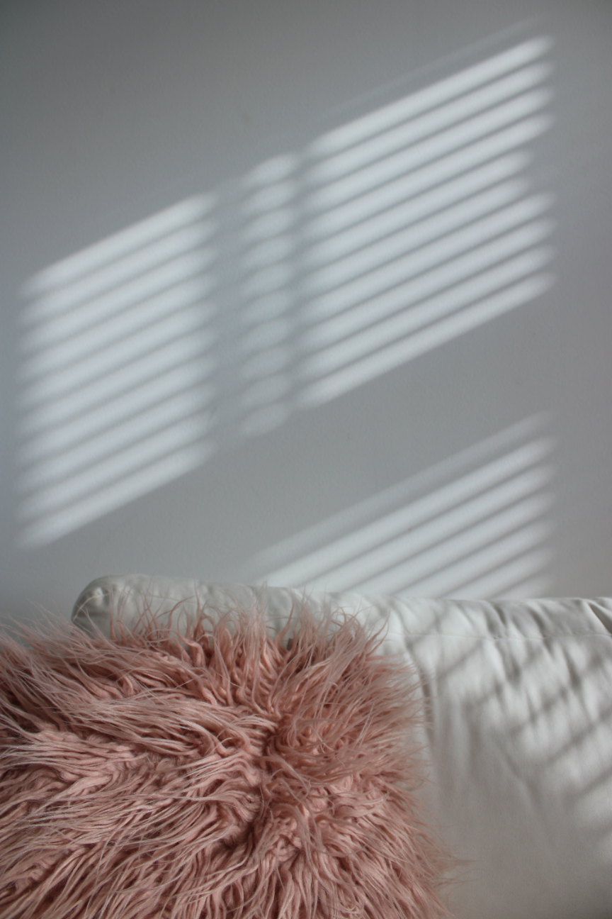 A pink furry cushion on a white bed with sunlight casting striped shadows on the wall.
