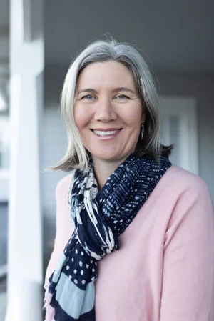 A smiling middle-aged woman with gray hair, wearing a pink sweater and a navy blue patterned scarf, standing outdoors near a house.