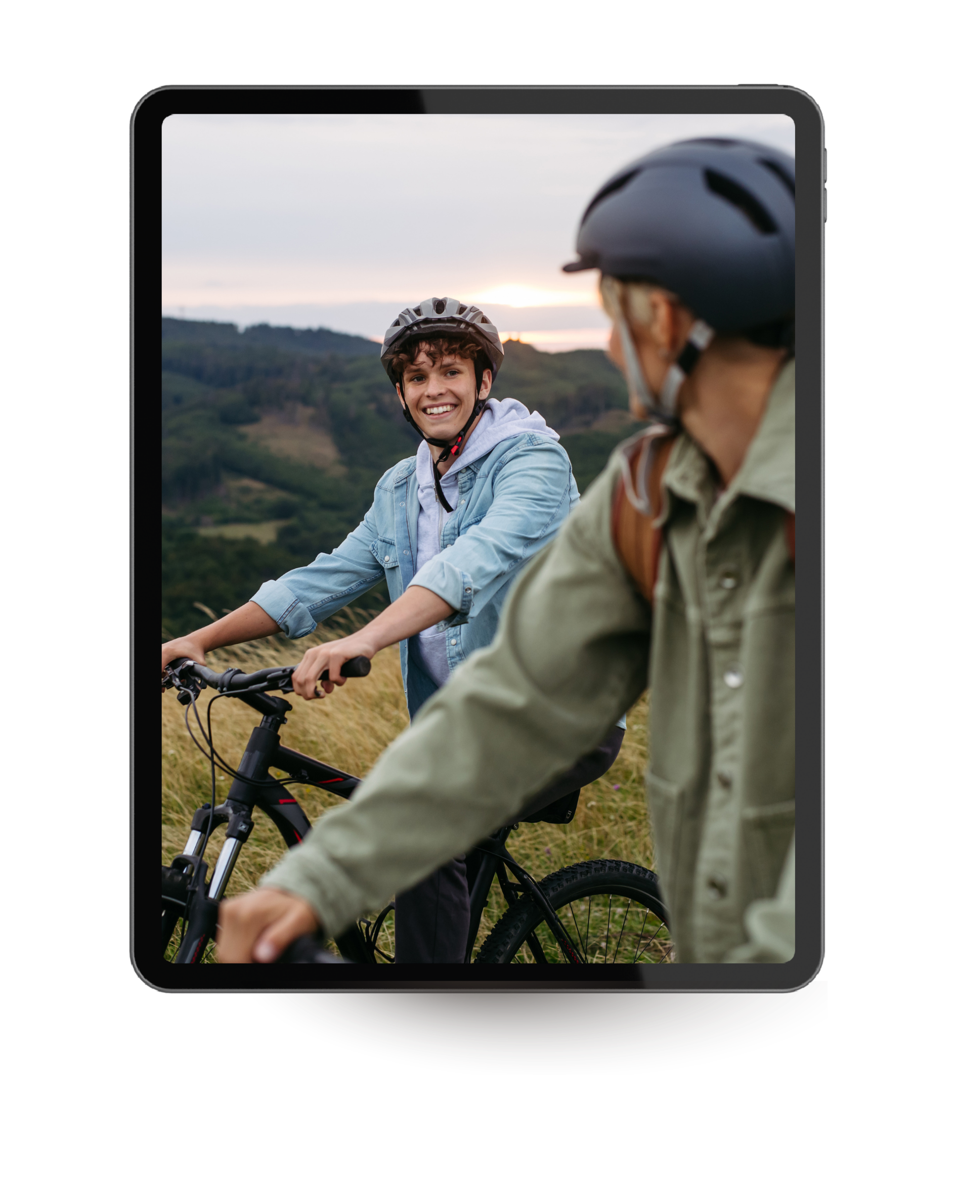 Two young men riding bikes on a grassy hill during sunset, wearing helmets and casual jackets, with mountains and a cloudy sky in the background.