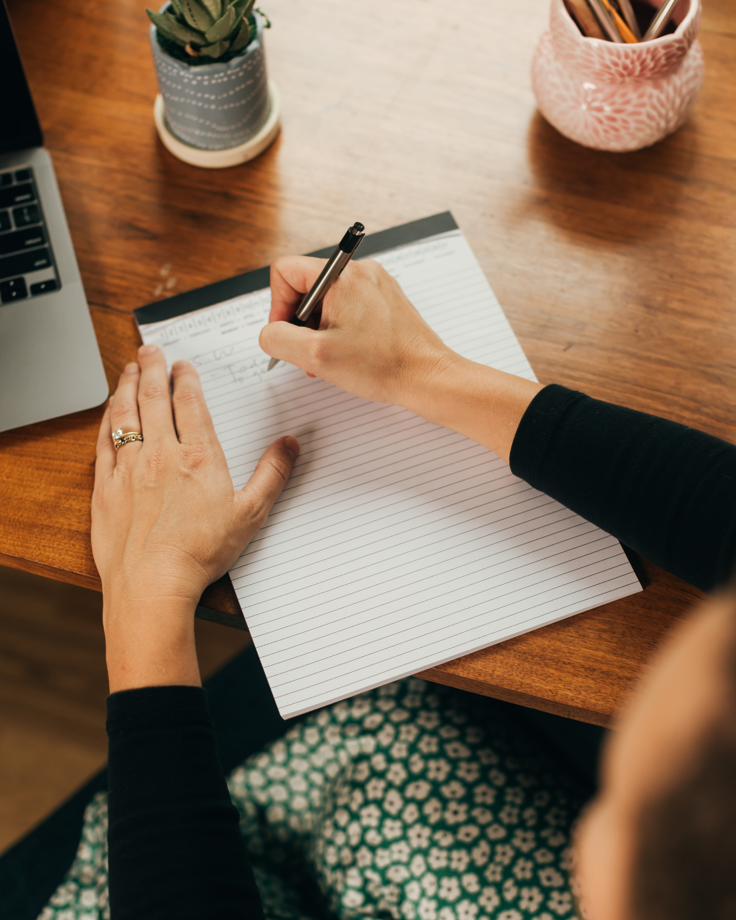 A person wearing a black long-sleeve shirt is writing in a spiral notebook on a wooden desk. The desk has a laptop on the left, a potted plant with a white and blue pot, and a pink patterned container holding pens and pencils. The person has a ring on their left hand and is dressed in patterned pants.