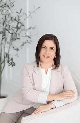 A woman with shoulder-length brown hair, smiling, sitting at a white table in a bright room with a potted plant in the background.