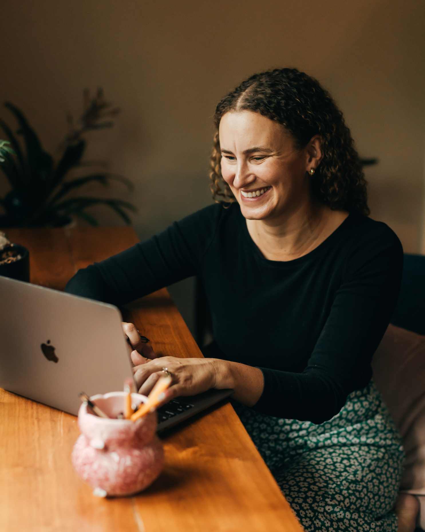 A woman with curly hair smiling while working on a laptop at a wooden table, with a plant in the background and a pink pencil holder in the foreground.