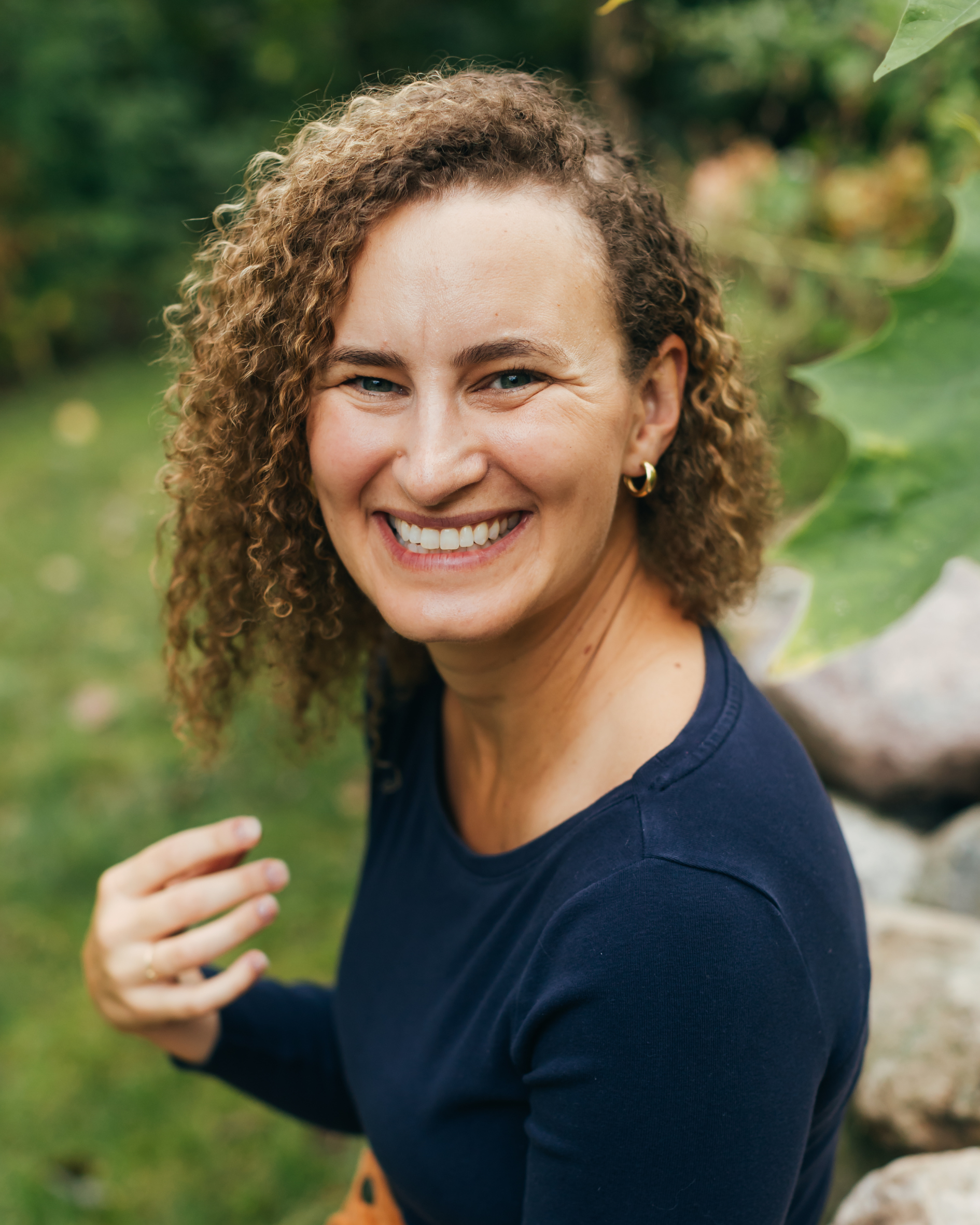 Close-up of a smiling woman with curly hair outdoors with greenery in the background.