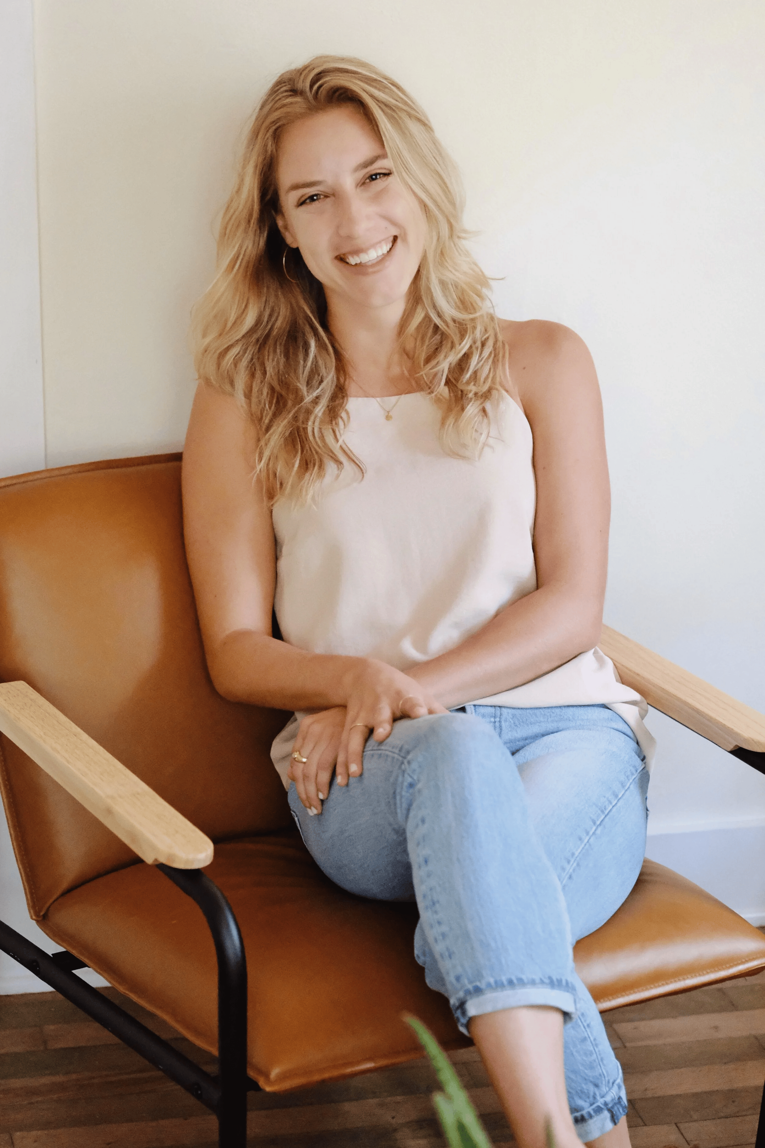 A woman with long, wavy blonde hair sitting on a brown leather chair with wooden armrests, smiling and looking at the camera, wearing a white tank top and light blue jeans.