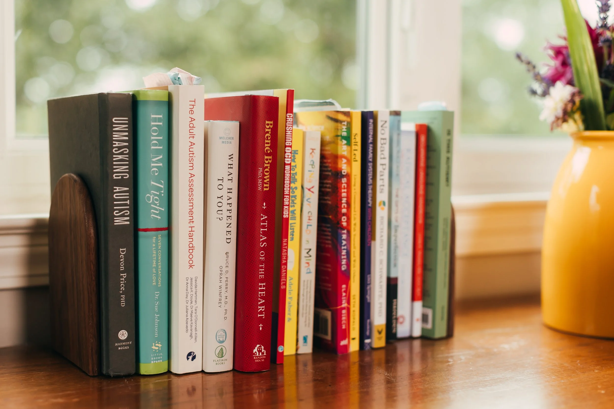 A row of colorful books on a wooden shelf near a window, with a blurred background of greenery outside and a yellow vase with flowers on the right.