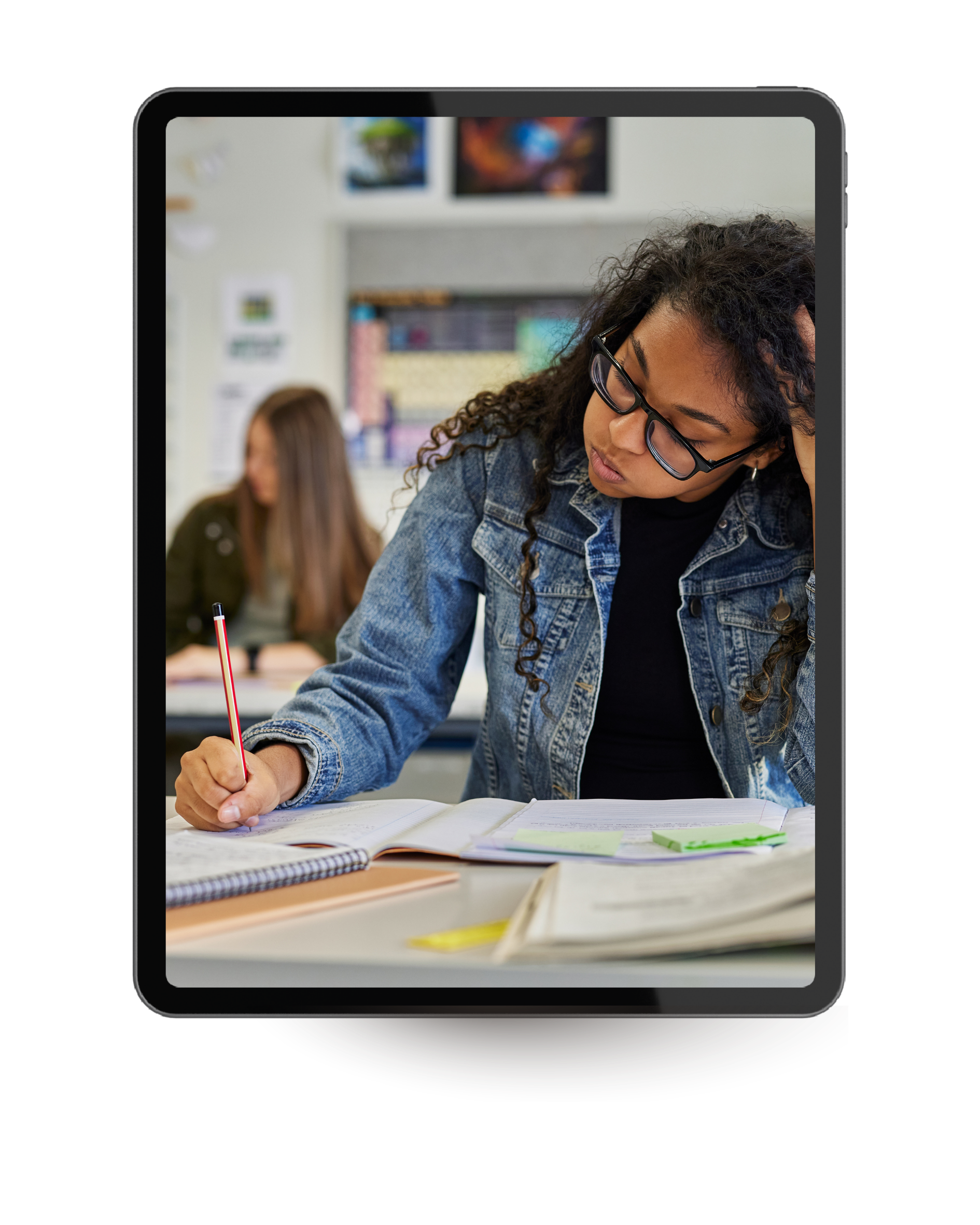 A young woman with curly hair and glasses is focused on writing in a notebook at a desk in a classroom. In the background, another girl is sitting and working, with classroom shelves and colorful artwork on the wall.