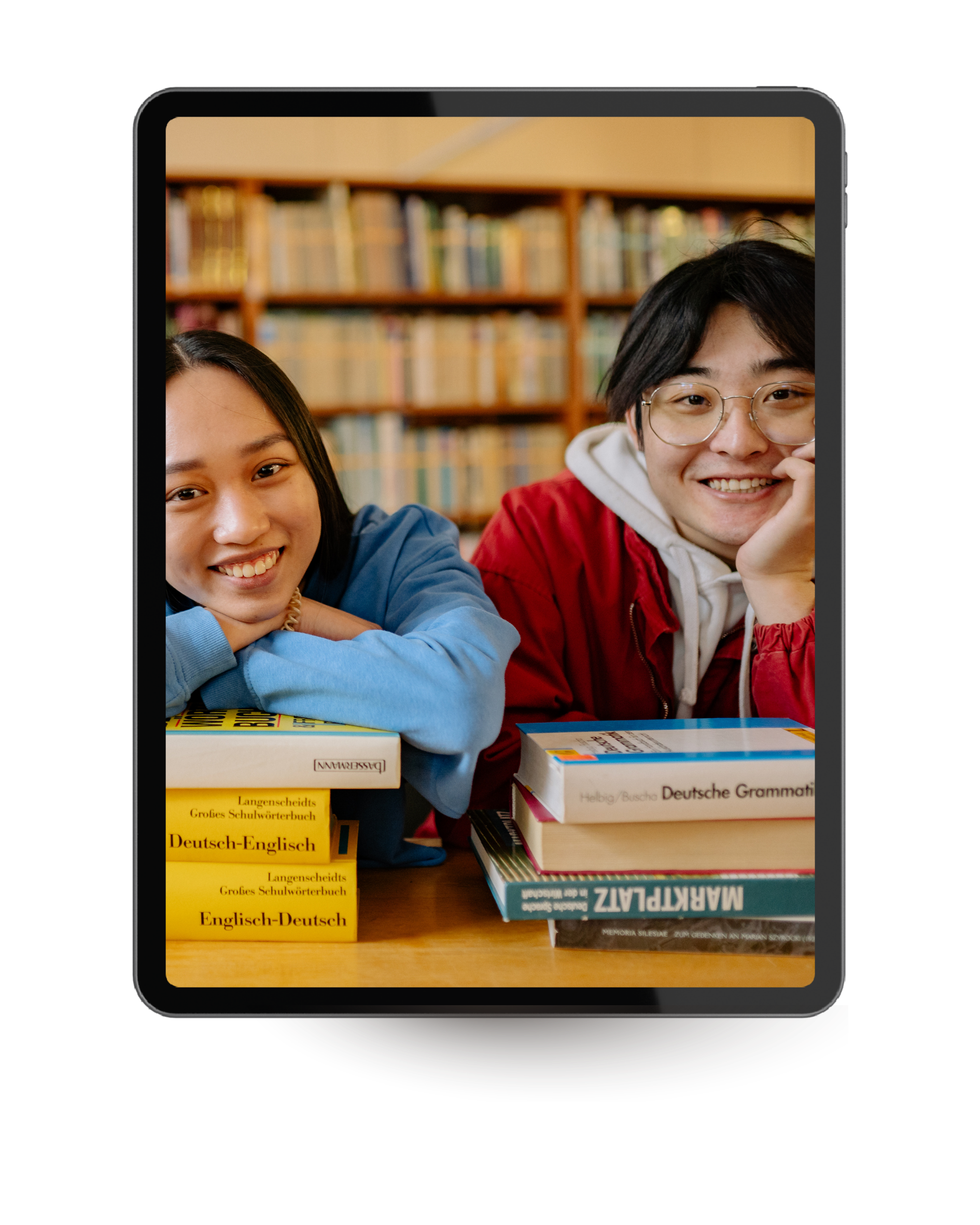 Two smiling students sitting at a table with stacks of German and English textbooks in a library or classroom.