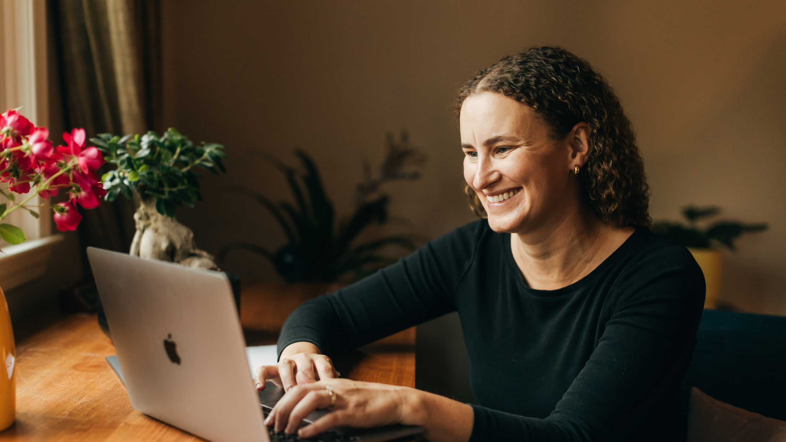 A woman with curly hair smiling while looking at her laptop in a cozy room with plants and flowers.