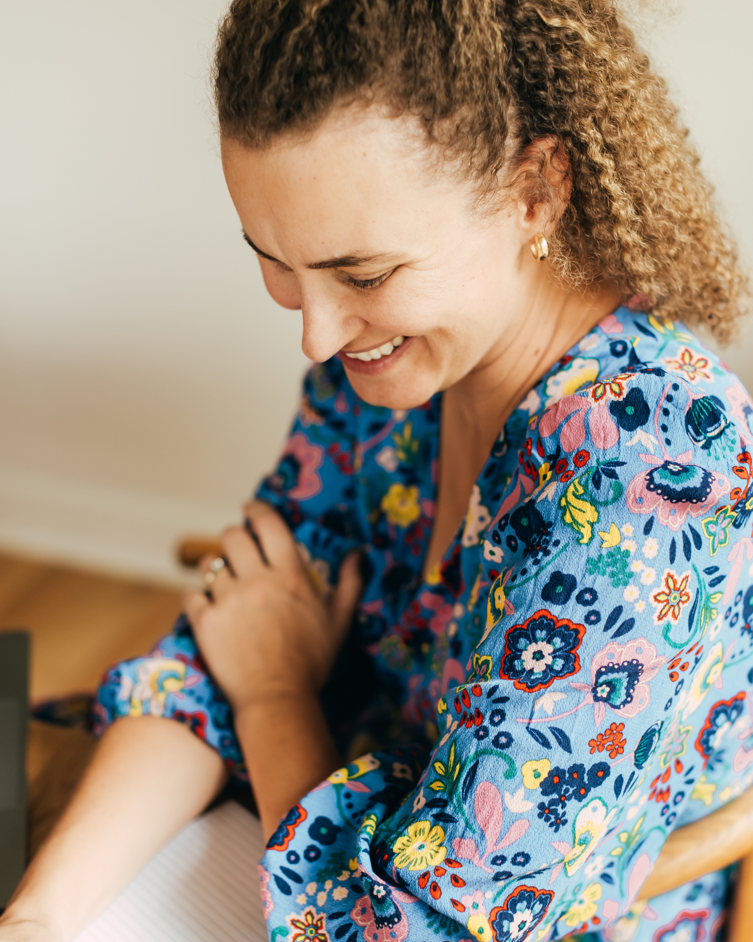 A woman with curly hair smiling and looking down, wearing a colorful floral blouse, sitting with her arm resting on a table.