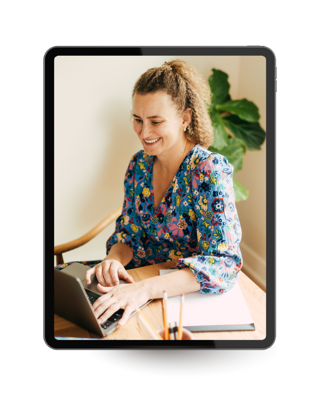 Woman with curly hair smiling and typing on a laptop at a wooden desk with a green plant in the background.