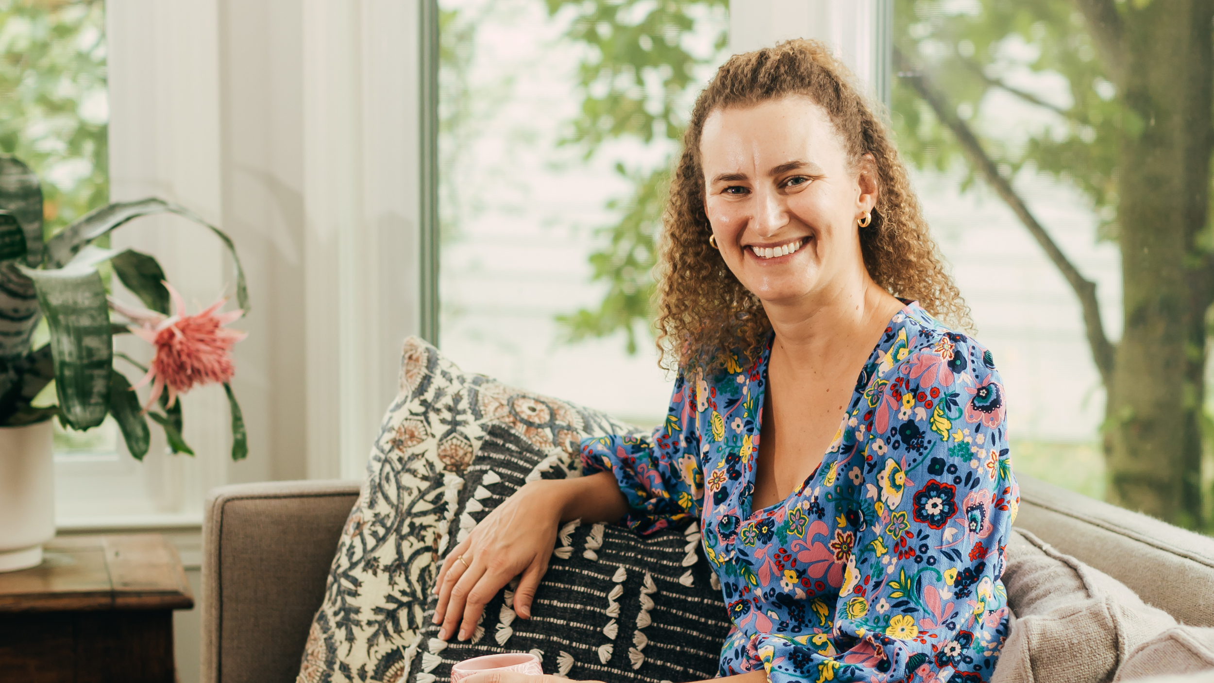 A woman with curly hair smiling while sitting on a sofa in a bright living room, wearing a colorful floral blouse. There are decorative pillows and a potted plant nearby, with large windows and greenery outside.