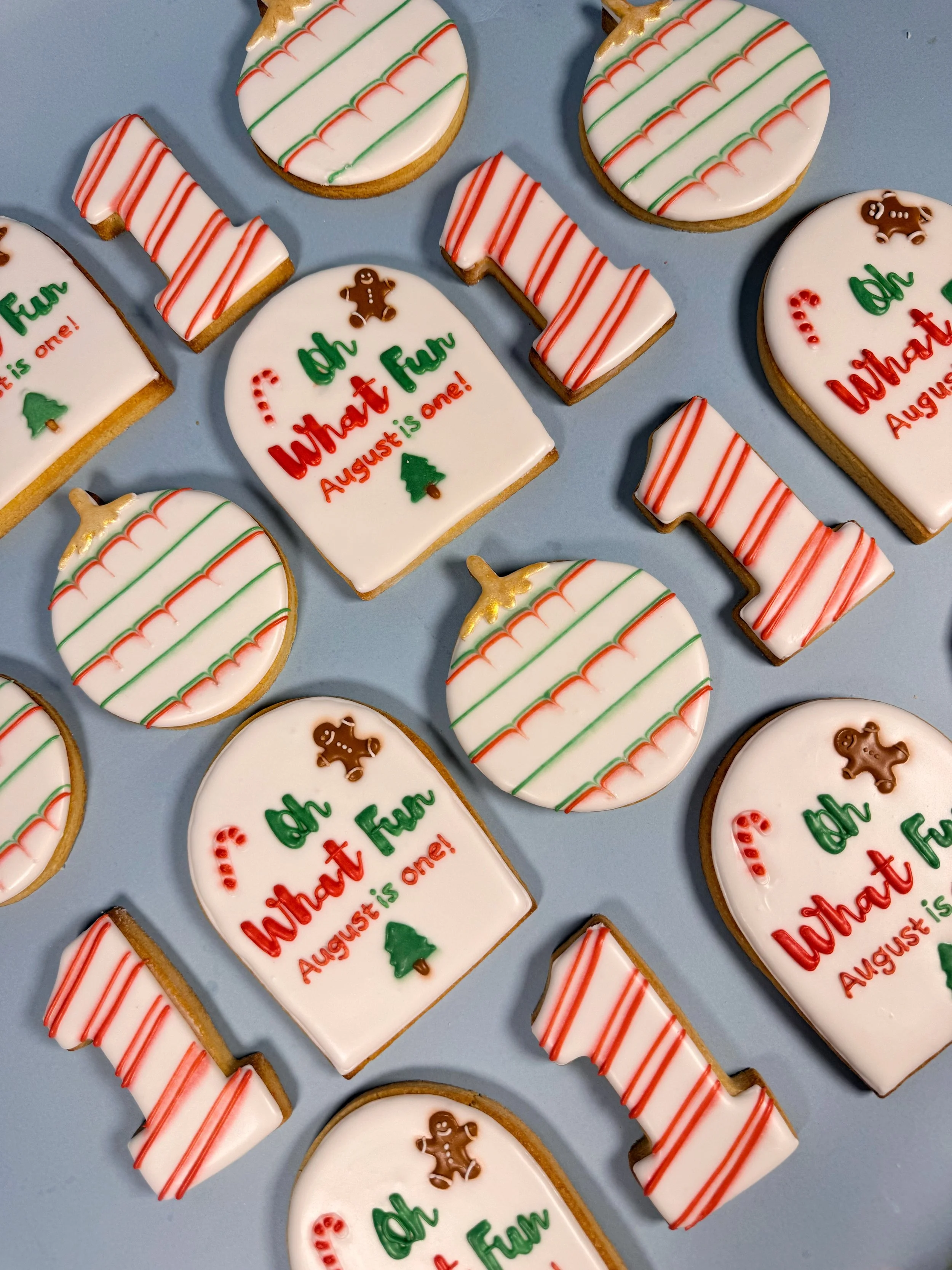 Decorated custom cookies themed for 1st birthday party with Christmas colors, featuring phrases like 'Oh What Fun' and 'August is one!', with gingerbread men, Christmas trees, and candy cane designs.