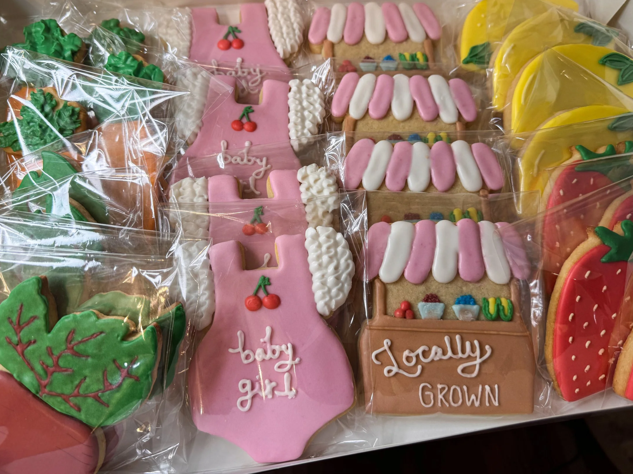 A variety of custom baby shower decorated cookies in a box, including some shaped like strawberries, carrots, a pink onesie with the word "baby" and cherries, and a small rectangular cake with flowers and the words "Locally Grown."