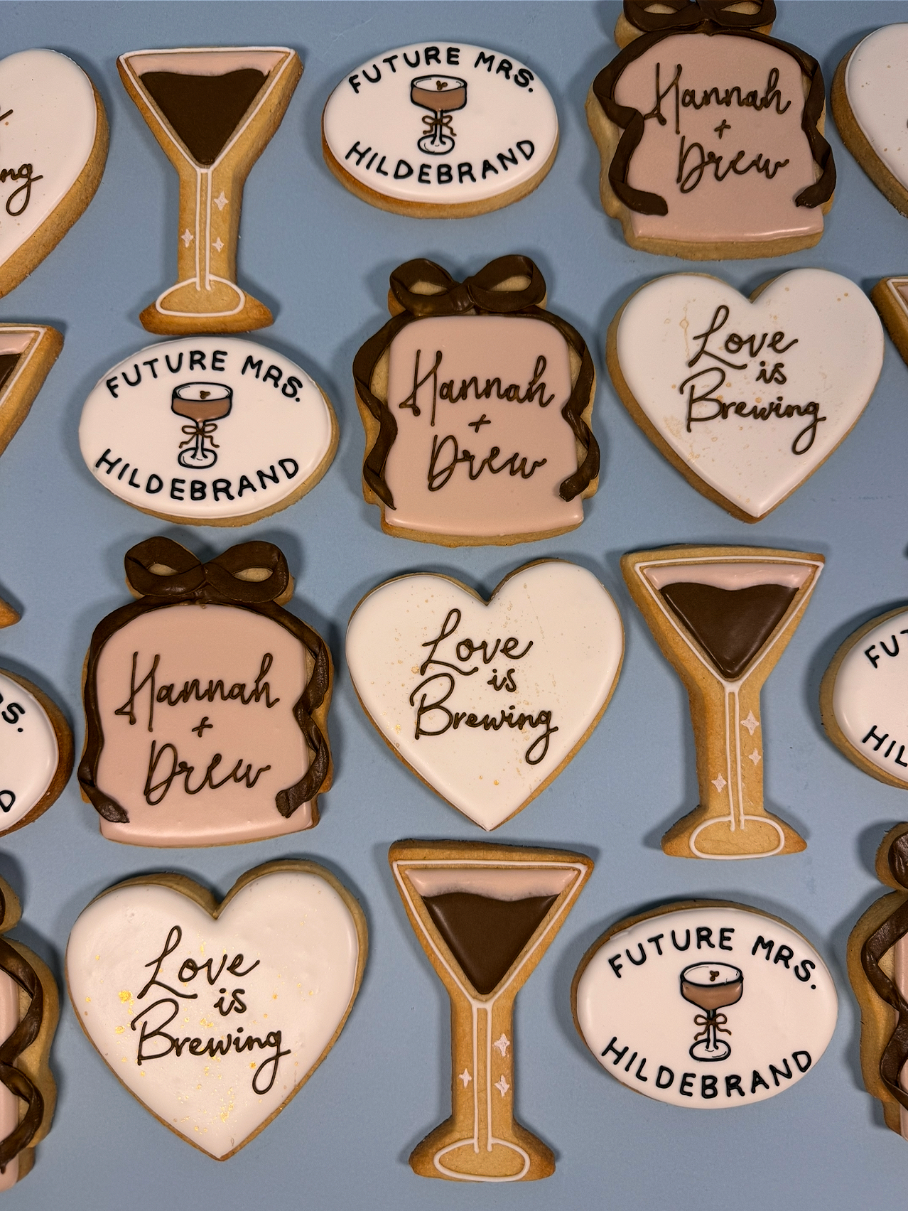 Decorative themed bridal shower cookies with various messages and designs, including glasses of champagne, hearts, and text such as "Future Mrs. Hildebrand," "Hannah + Drew," and "Love is Brewing."