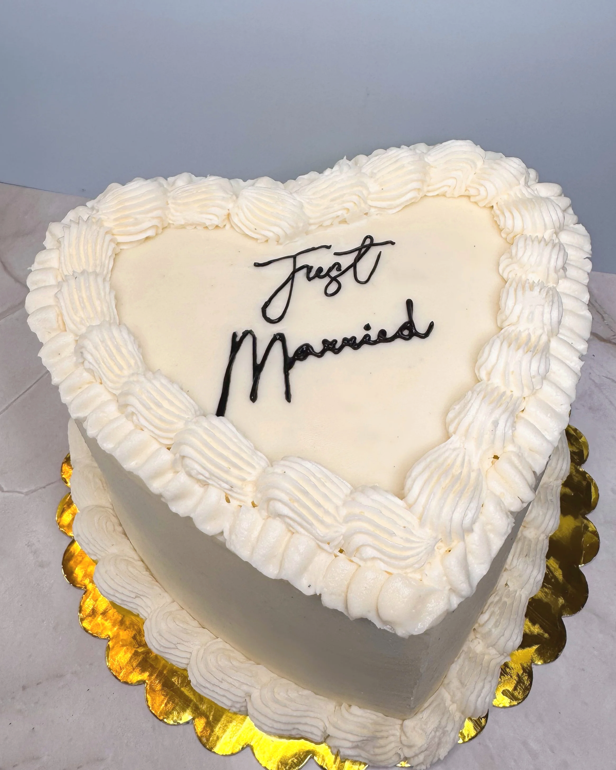A heart-shaped wedding cake with white frosting and piped border, with the words 'Just Married' written in black icing on top.
