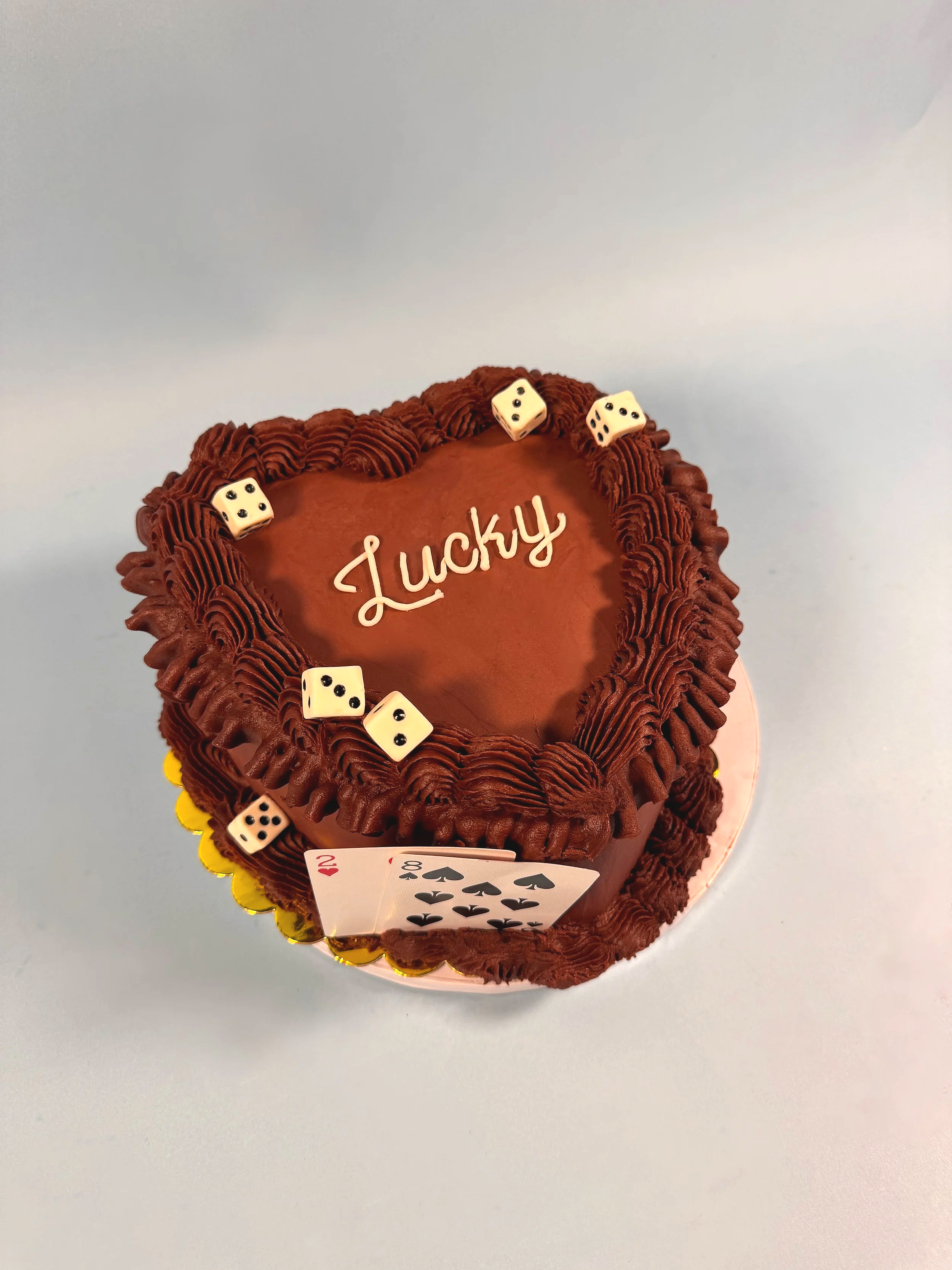 A heart-shaped burgundy birthday cake decorated with dice and playing cards, with the word 'Lucky' written on top.