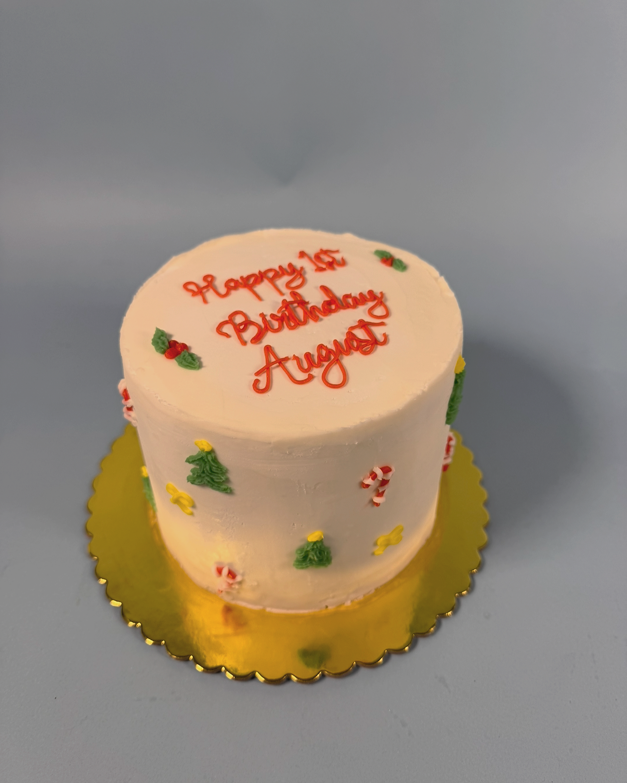 White birthday cake with holly, Christmas trees, and candy cane decorations on a gold tray, with red writing that says 'Happy 1st Birthday August'.