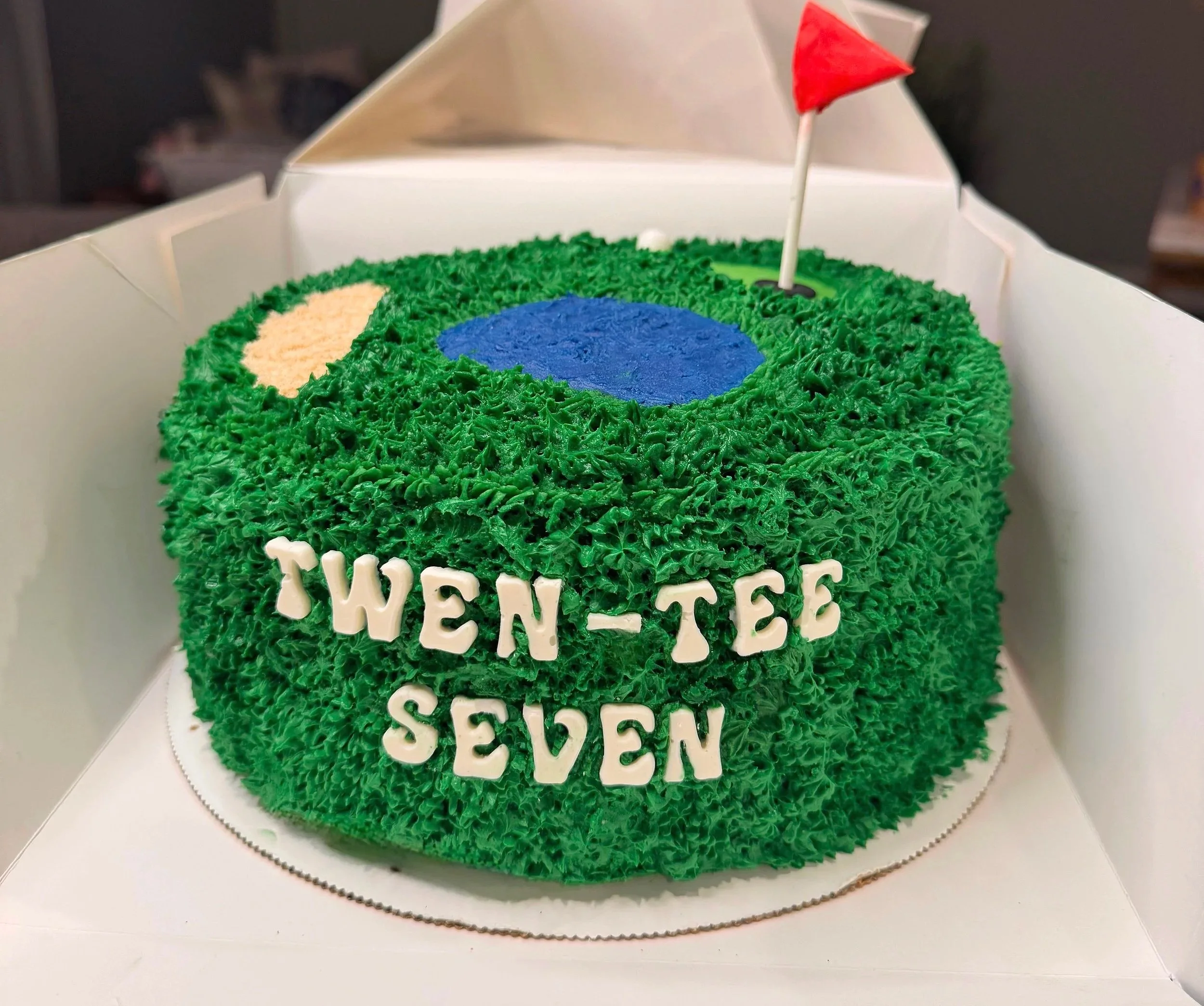 A birthday cake decorated to look like a golf course with grass-like green frosting, a blue pond, a sand trap, a red flag on a golf hole, and white spelling out "Twen-tee Seven" on the side.