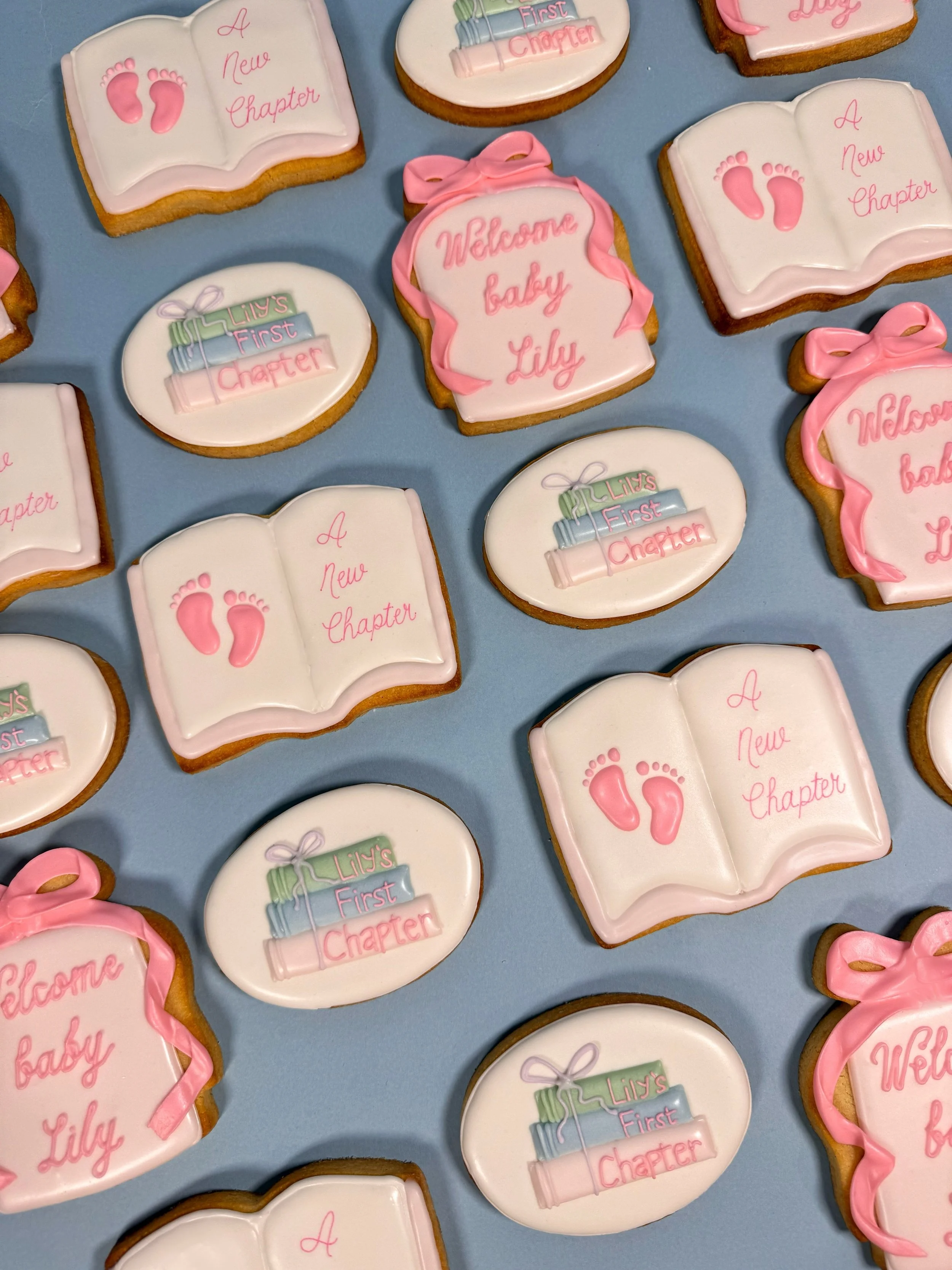 Decorative custom baby shower cookies with pink, white, and pastel-colored icing, featuring messages like "Welcome baby Lily," "A new chapter," and illustrations of baby footprints, books, and a baby rattle.