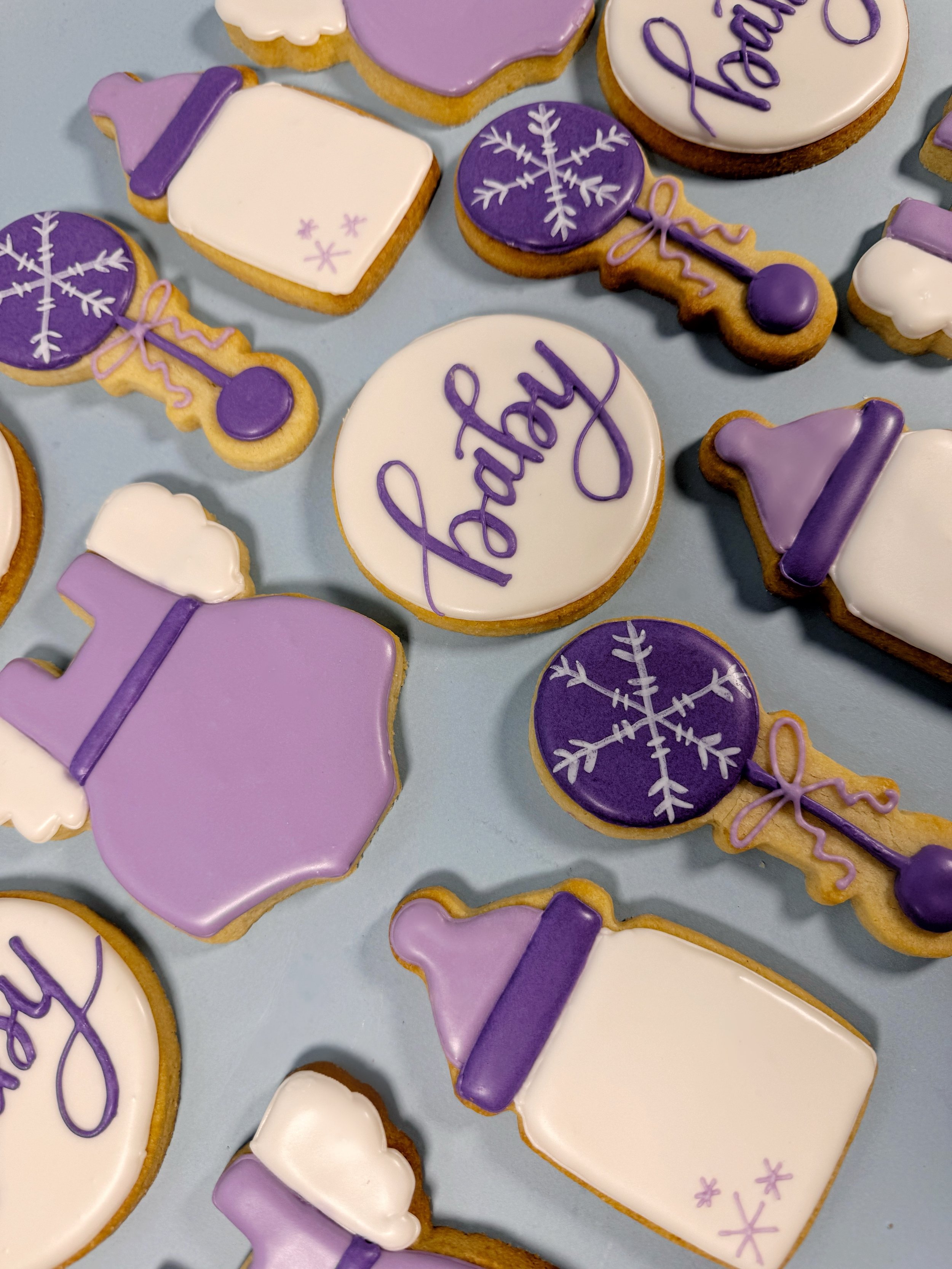 Collection of decorated cookies with purple, white, and lavender icing, featuring snowflakes, baby bottles, and the word 'baby' written on one cookie.