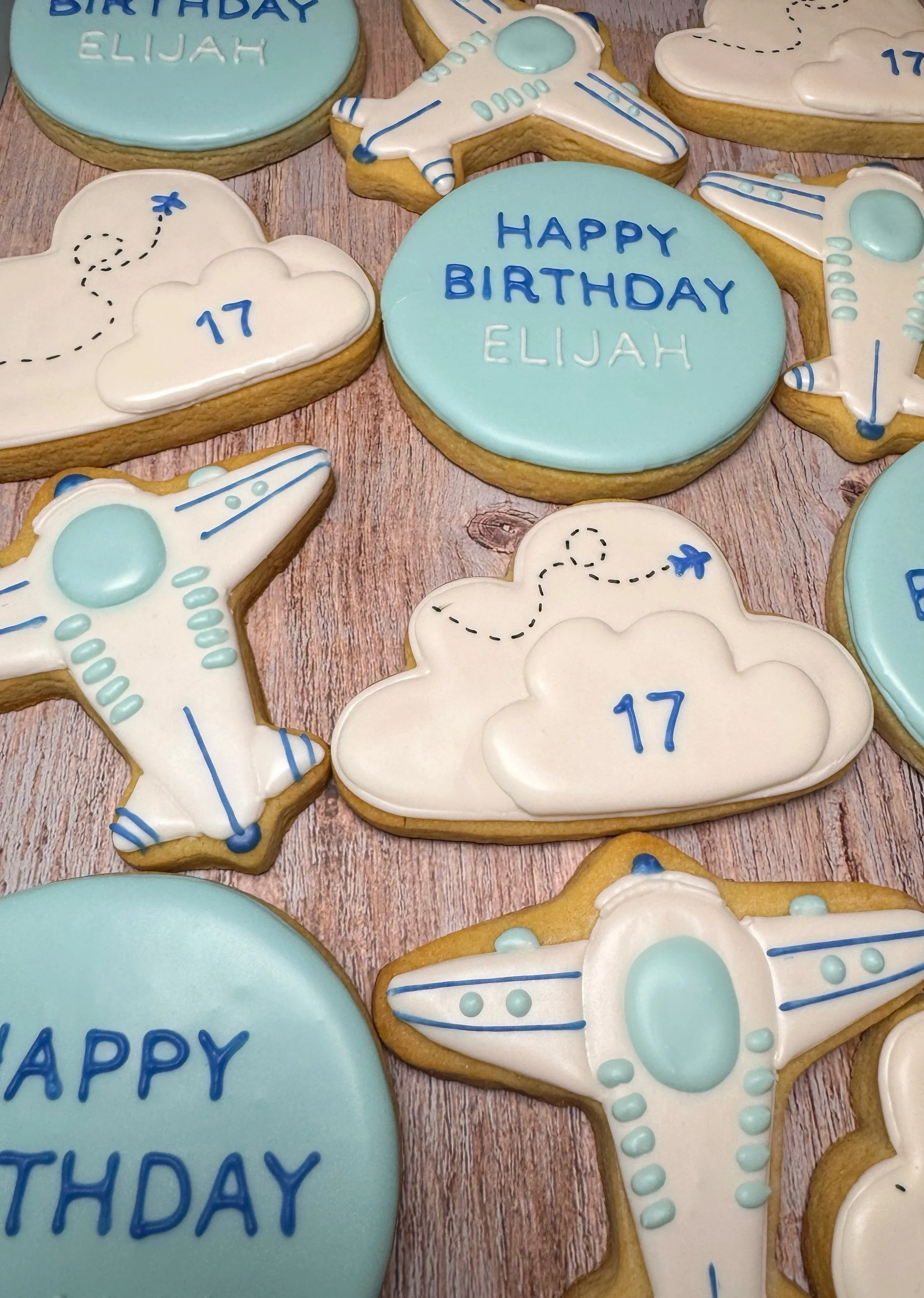 Cookies with space-themed designs, including rockets, clouds, and planets. Some cookies have messages like "HAPPY BIRTHDAY ELIJAH" and the number 17, decorated in blue, white, and light blue icing.