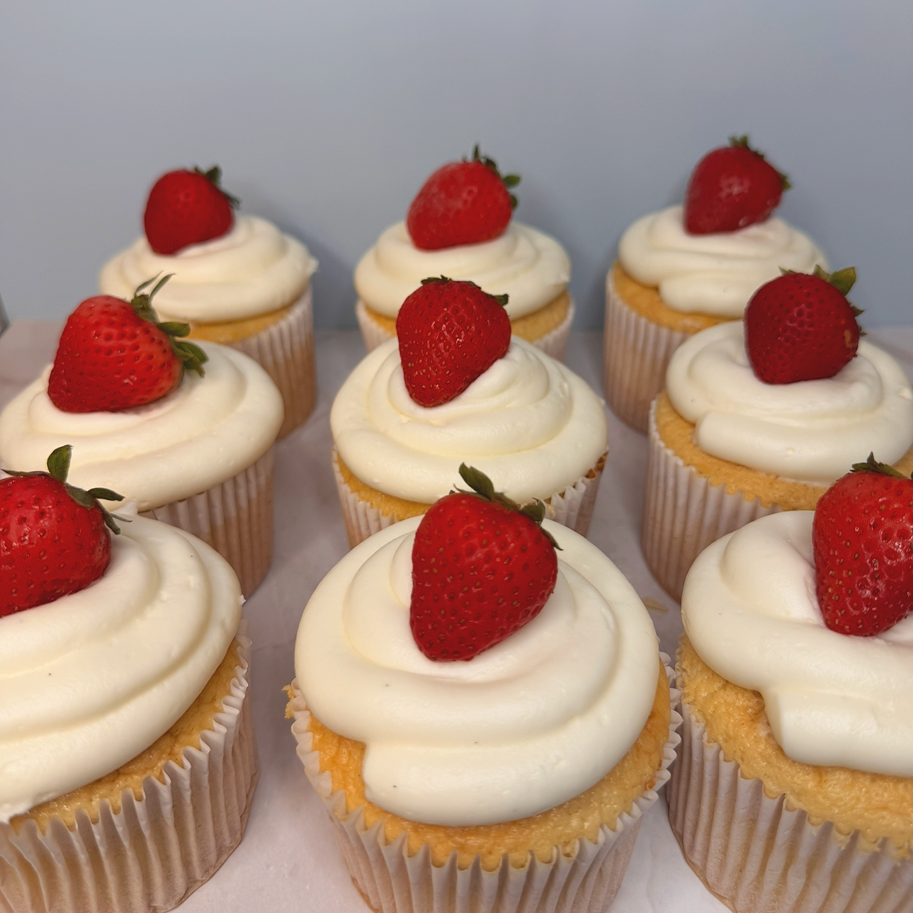 Nine vanilla cupcakes topped with white frosting and a single fresh strawberry on top, arranged in three rows of three.