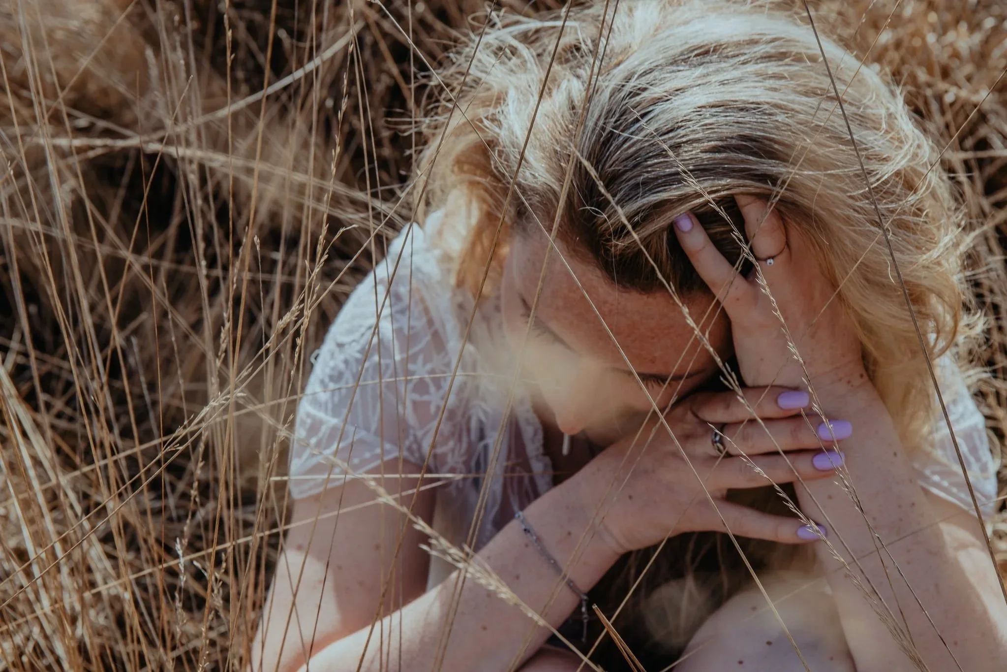 Vrouw met blond haar ligt in gras met haar gezicht naar beneden, haar handen op haar hoofd en gezicht.