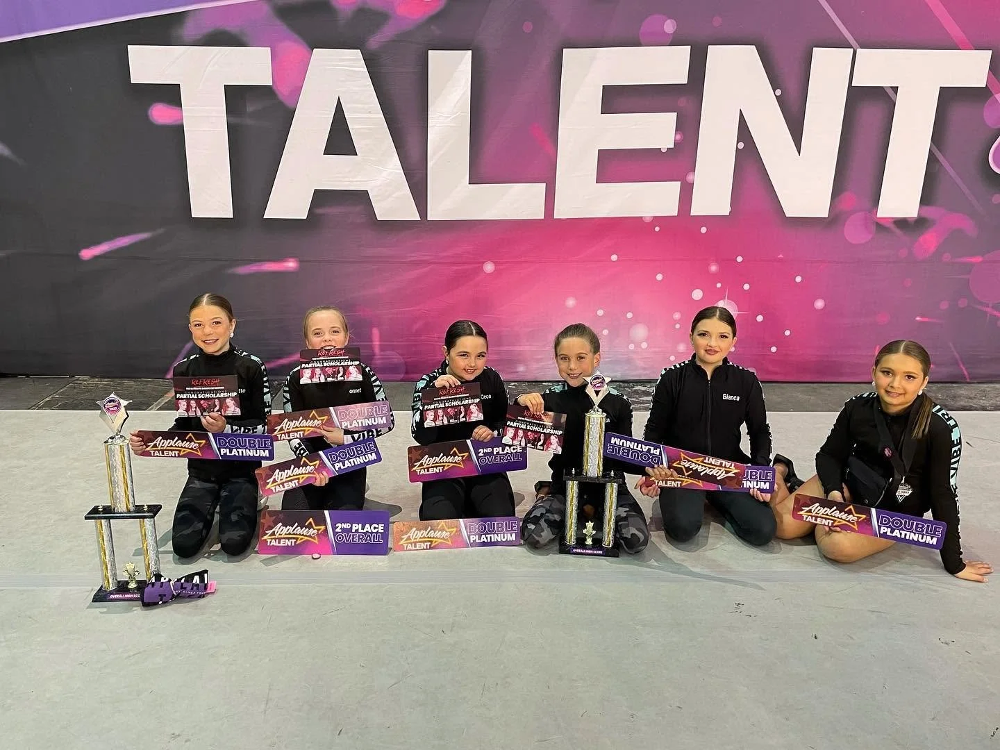 Six young girls in black dance costumes with awards and trophies, posing on a stage with a large pink and purple "TALENT" backdrop.