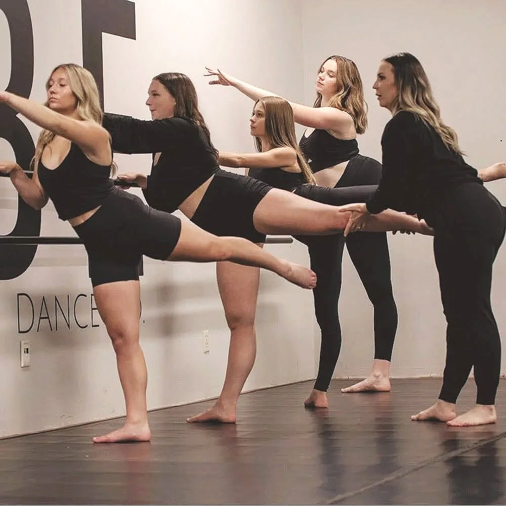 Five women in black workout clothes are practicing an exercise on a dance studio wall, with their arms extended forward and one leg lifted behind them, balancing on one foot.