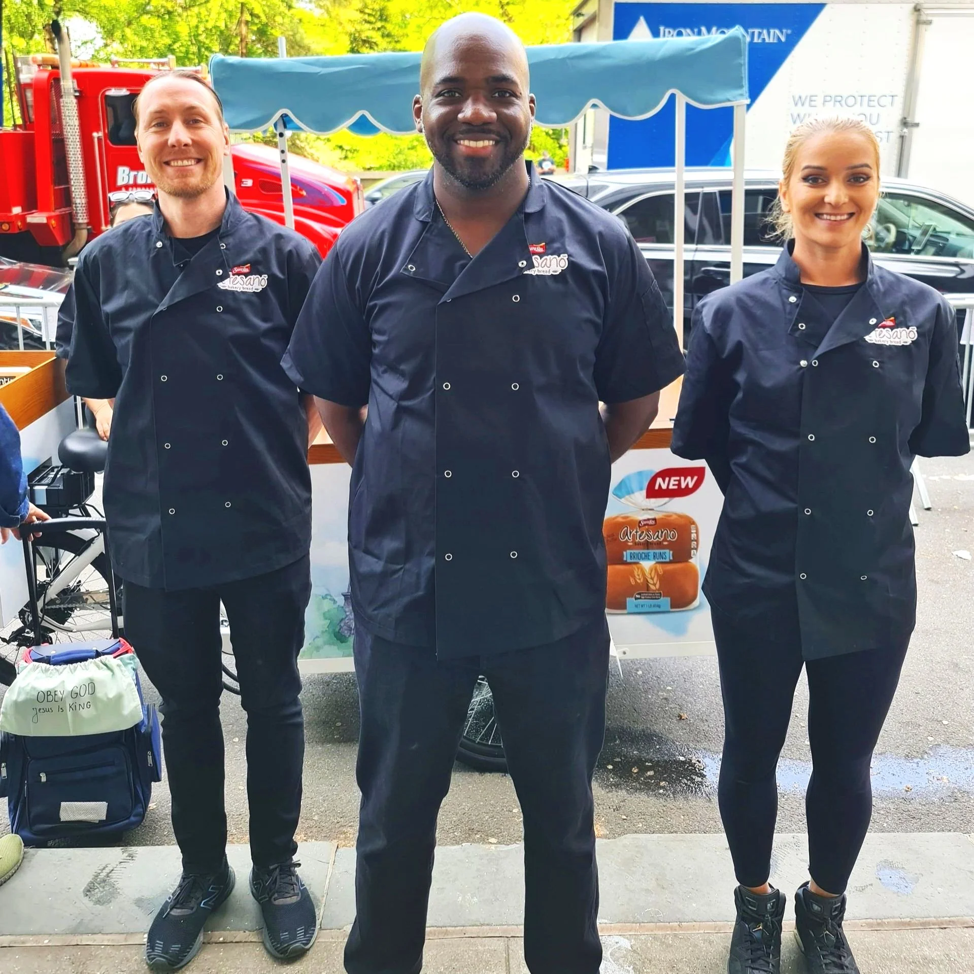 Three chefs in black uniforms standing outdoors in front of a food stall with a red truck and blue-and-white delivery truck in the background, smiling as they showcase a product labeled 'NEW.'