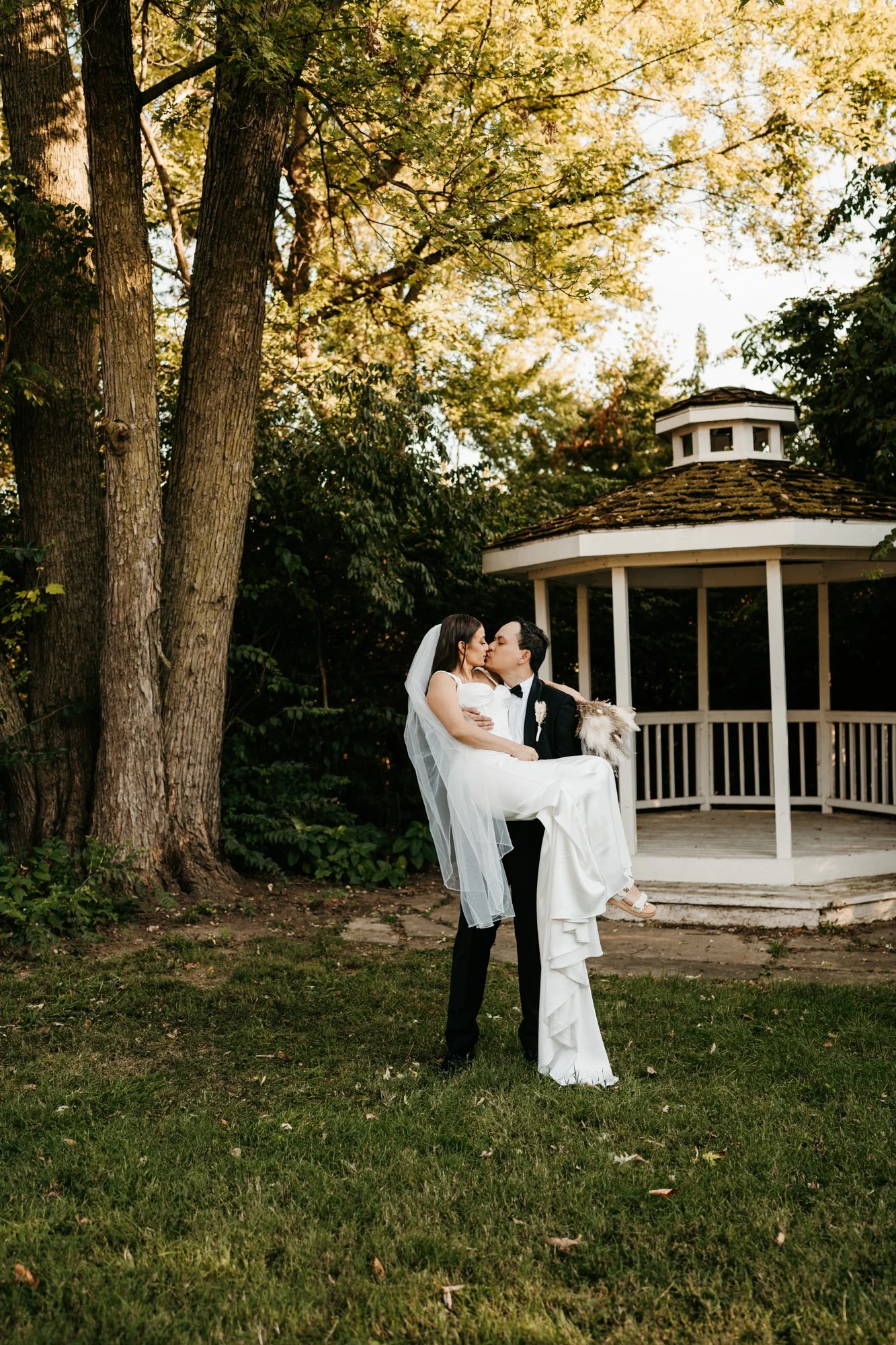 Bride and groom share a kiss outdoors during their wedding, with trees and a gazebo in the background.