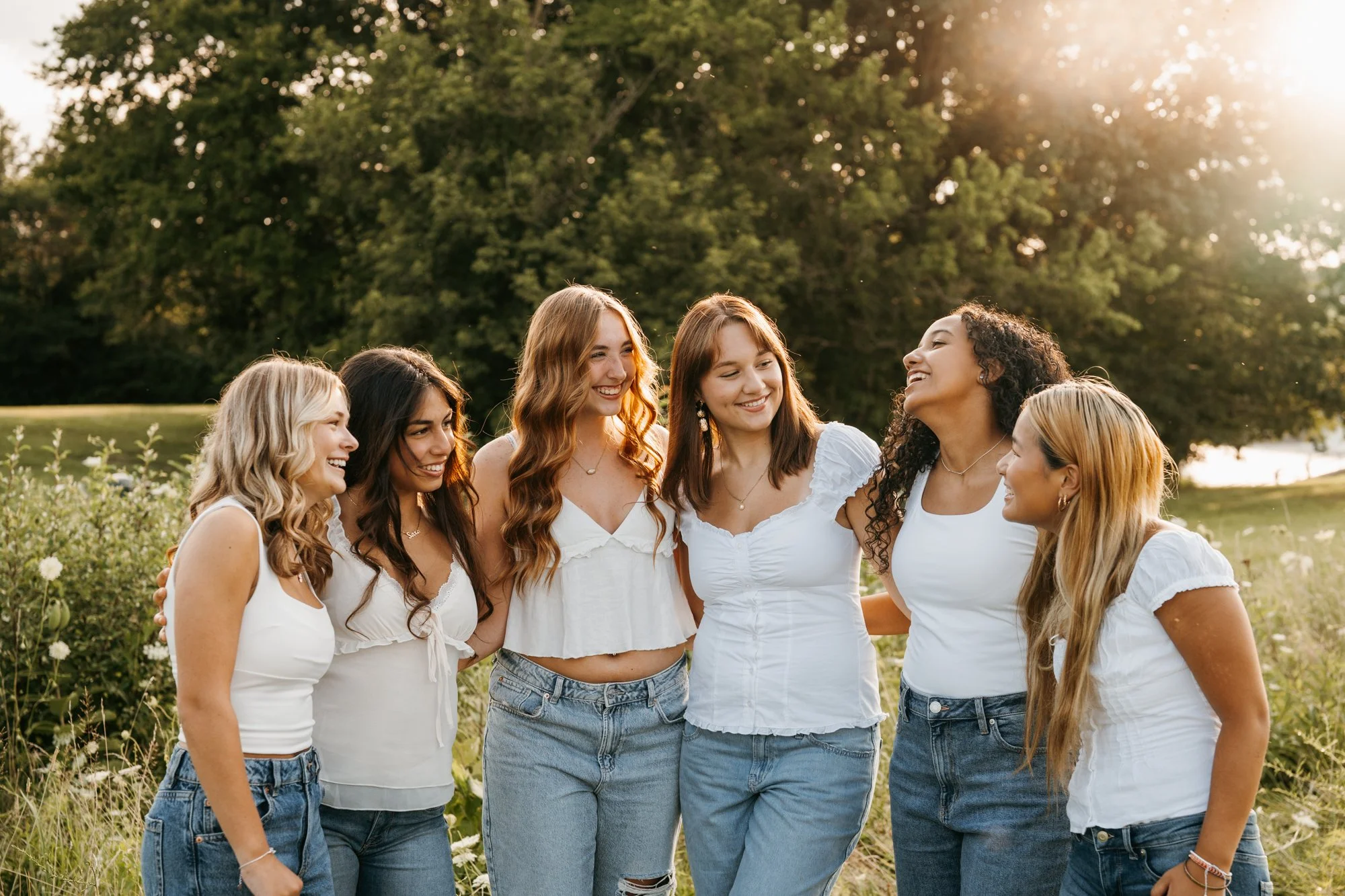 Six young women standing together outdoors in a field with greenery, smiling and talking, during the golden hour sunlight.