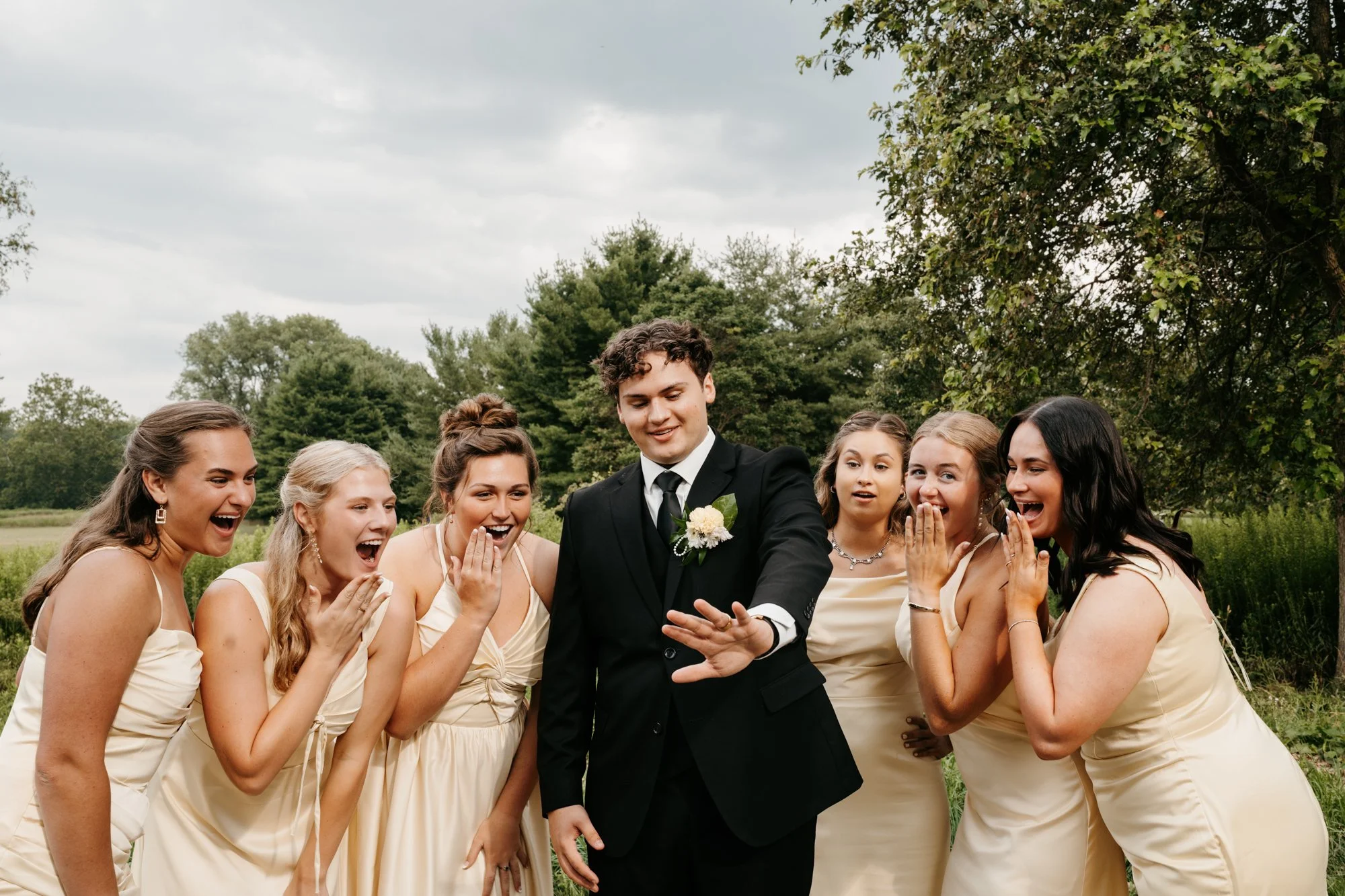 Group of young women in cream-colored dresses and a young man in a black suit with a boutonniere, outdoors against trees and a cloudy sky. They are smiling and reacting with surprise and excitement.