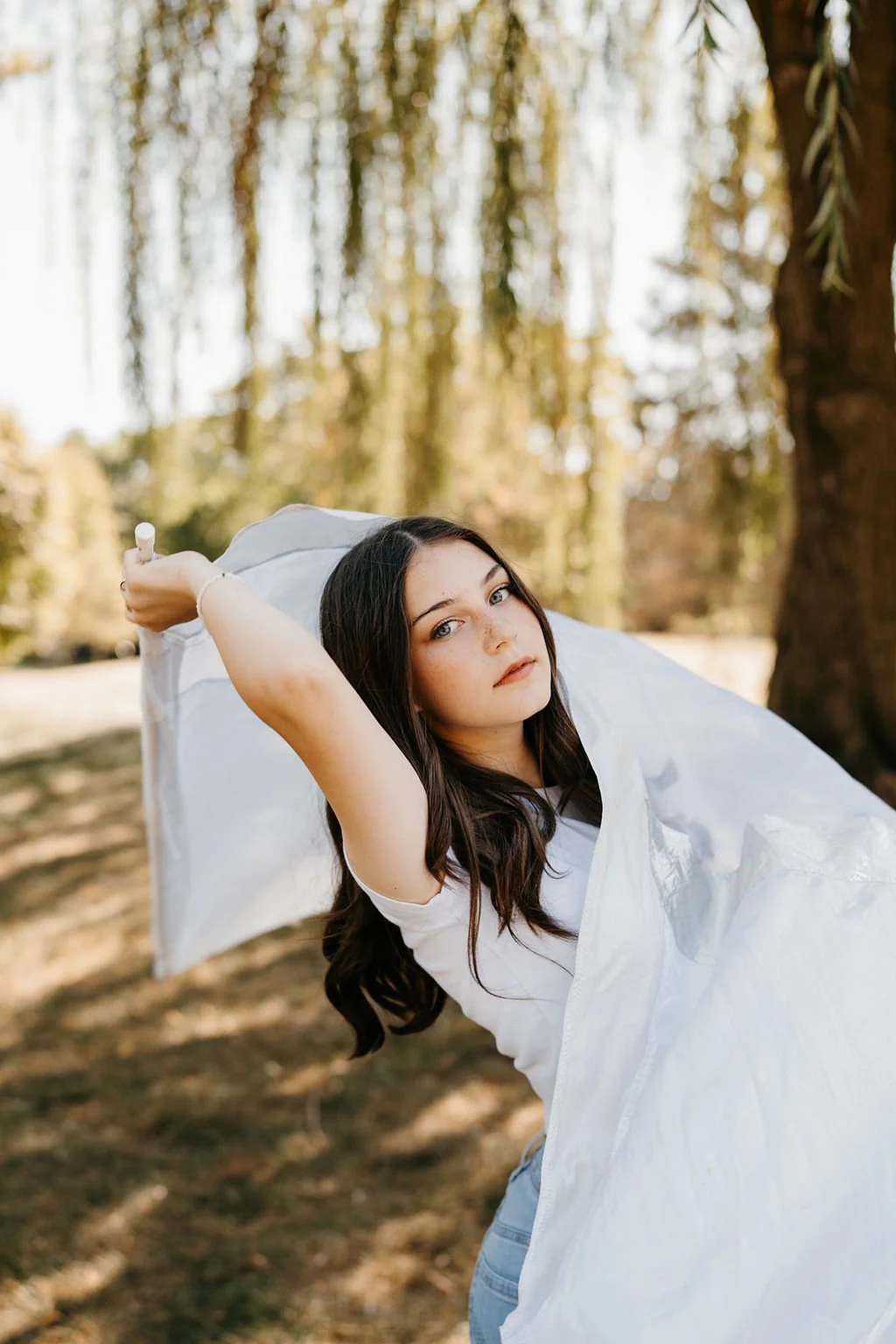 A young woman outdoors holding a white plastic bag, sunlight filtering through trees, she has long dark hair and a serious expression.