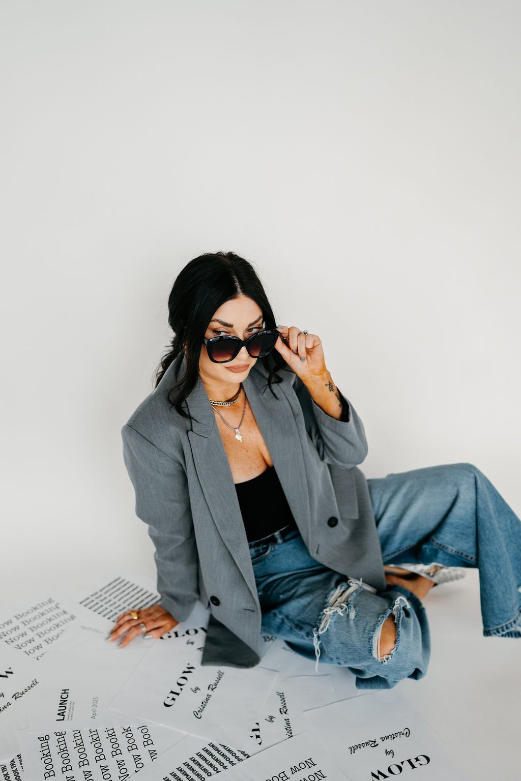 A woman with black hair wearing a gray blazer, black top, ripped jeans, and sunglasses seated on the floor surrounded by printed papers.