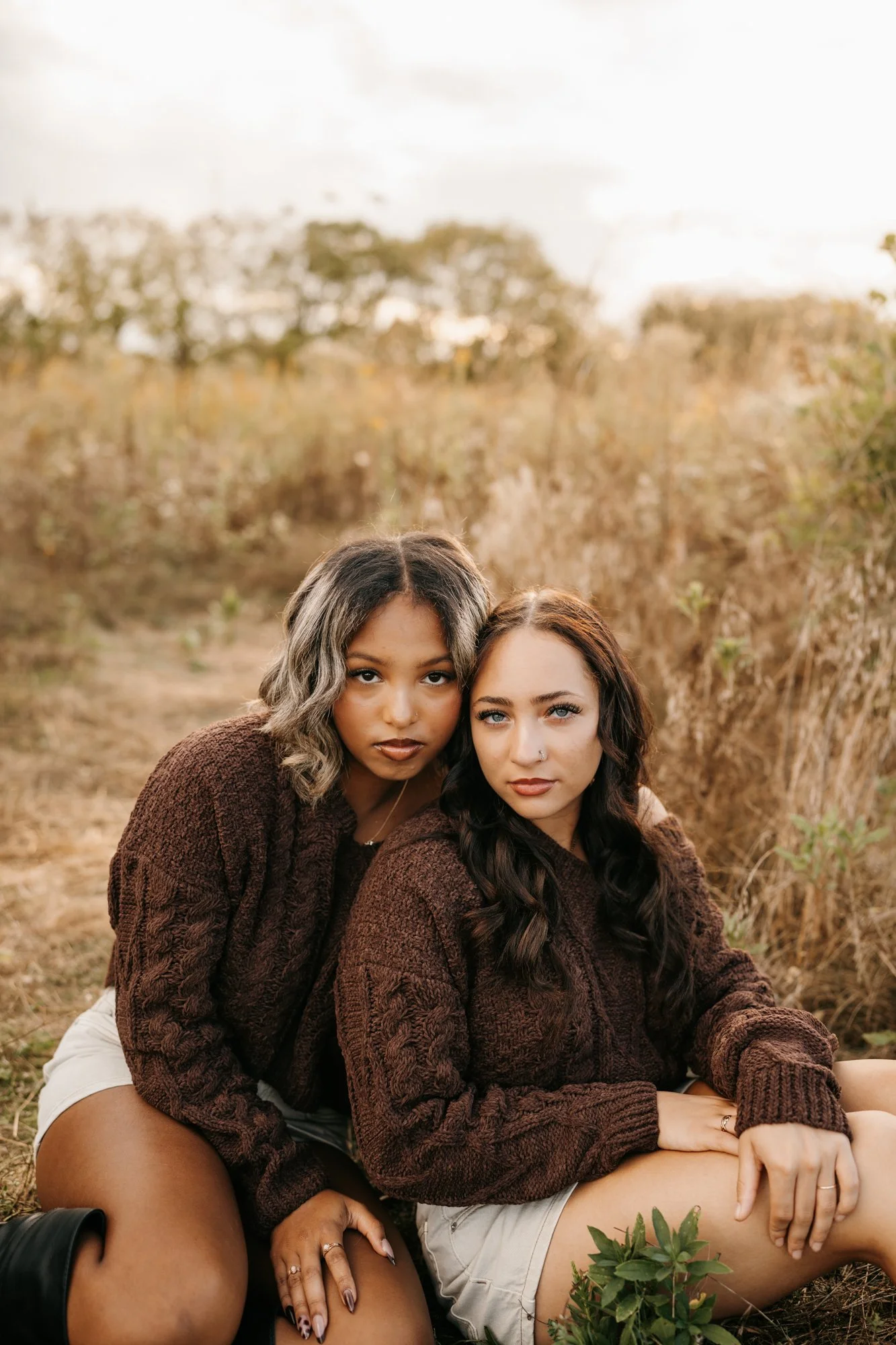 Two women sitting closely outdoors in a field with tall grass, wearing brown sweaters and light-colored shorts, looking directly at the camera.