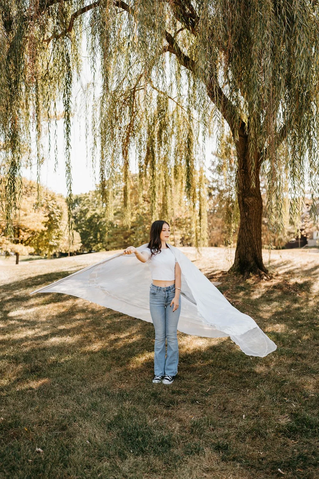 A young woman standing outdoors on a grassy area, holding a large white sheet or fabric behind her, with a large weeping willow tree overhead and autumn-colored trees in the background.