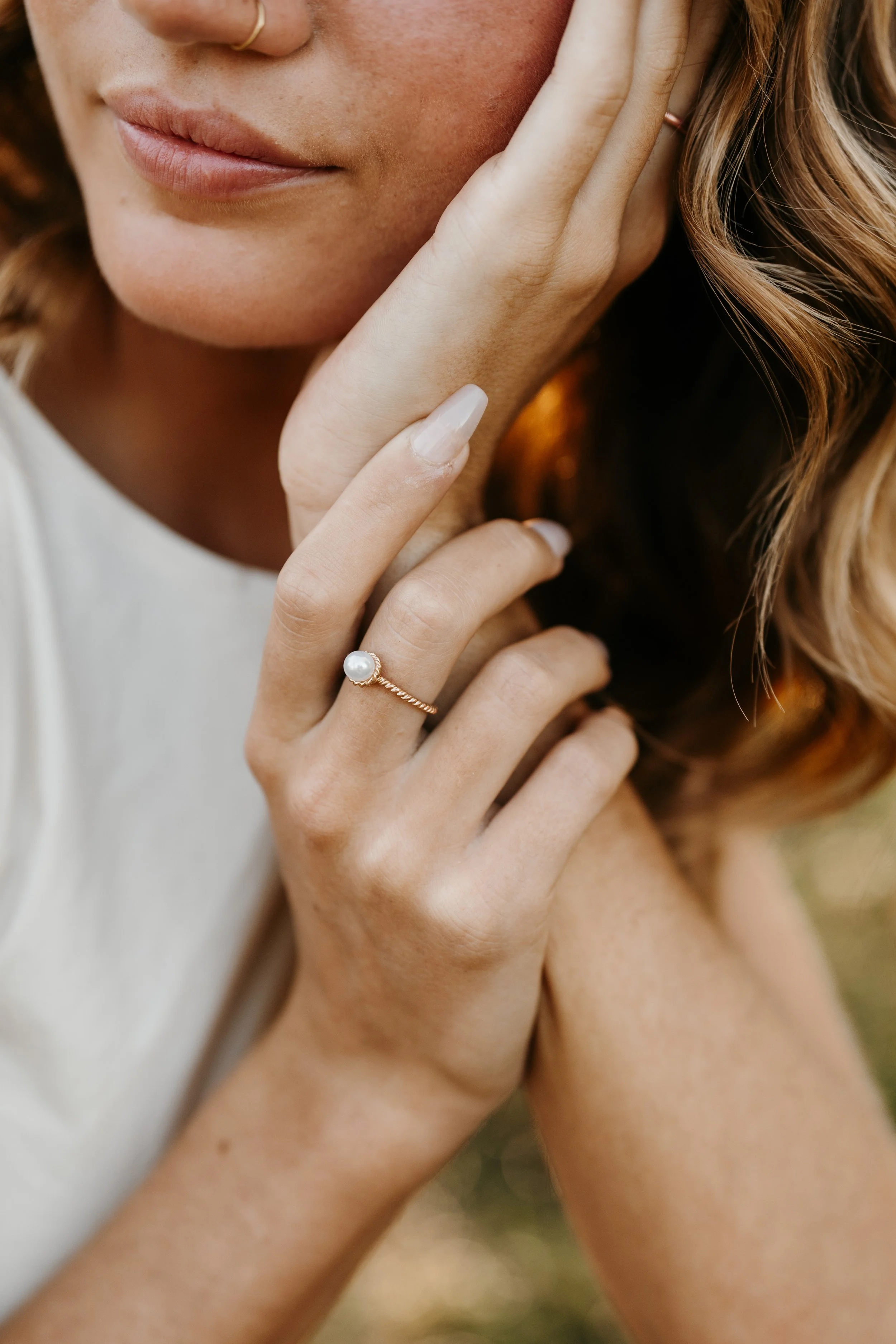 Close-up of a woman's face and hands with a pearl ring on her finger, touching her cheek, with wavy auburn hair and natural makeup.