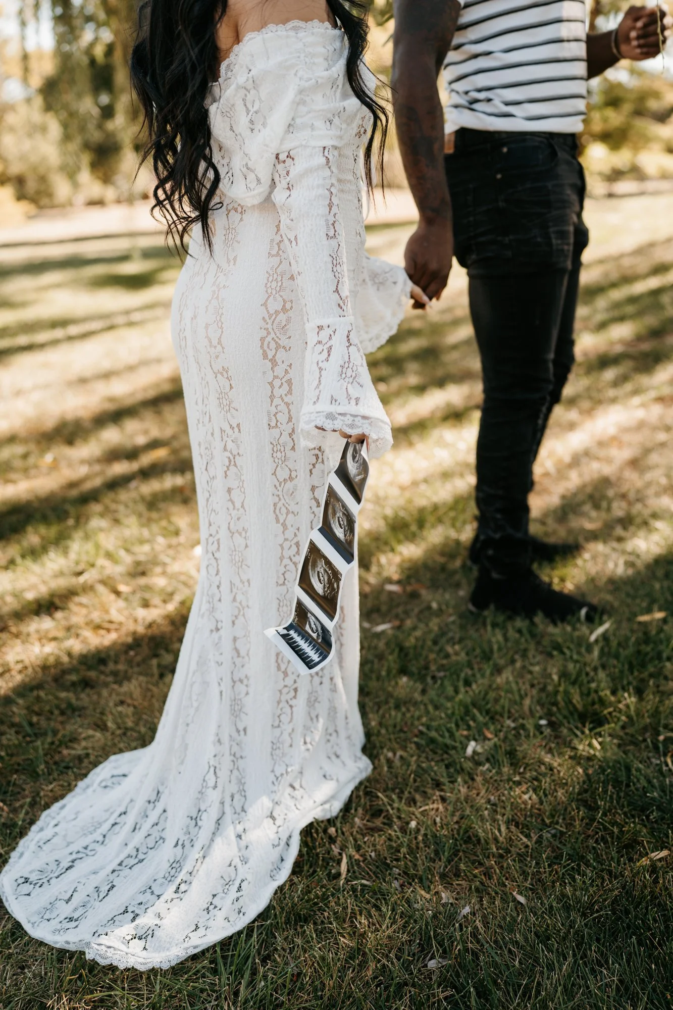 A woman in a white lace dress holding ultrasound images outside, with another person in a striped shirt holding her hand, on a grassy area.