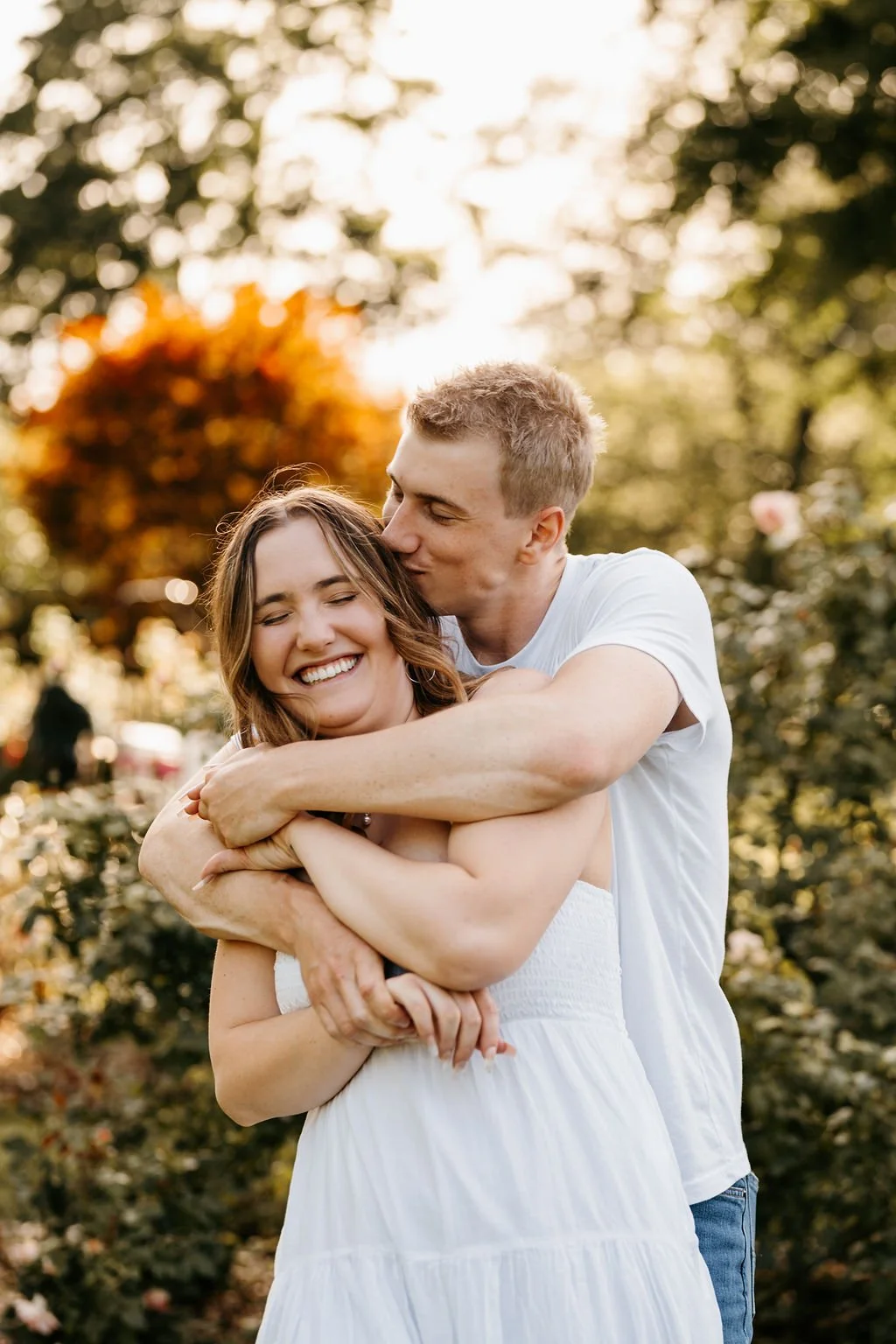 A man kisses a woman on the forehead as they share a hug outdoors during sunset, surrounded by trees with autumn foliage.