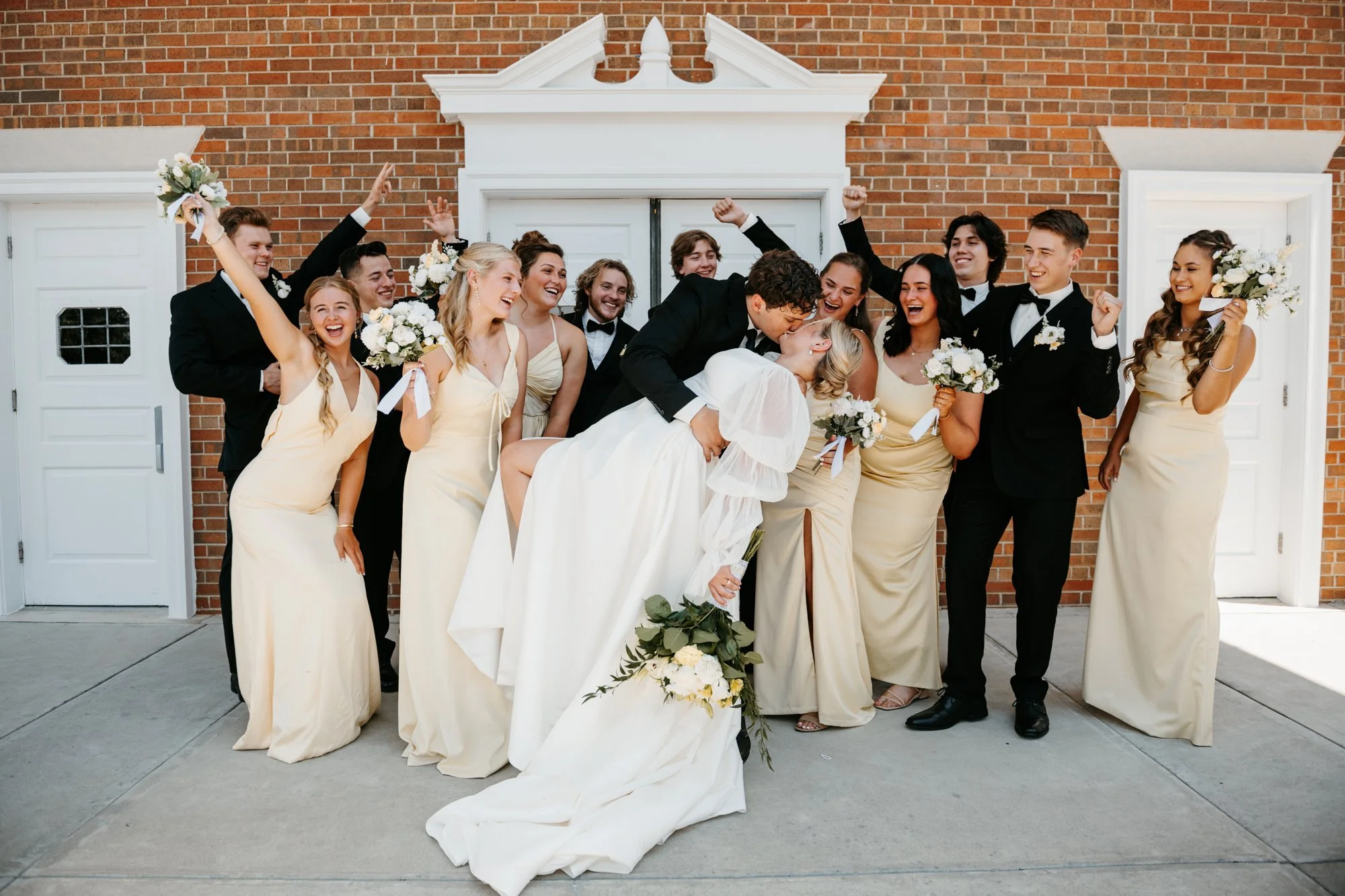 A wedding party in front of a brick building, featuring the bride and groom kissing, surrounded by bridesmaids and groomsmen celebrating.