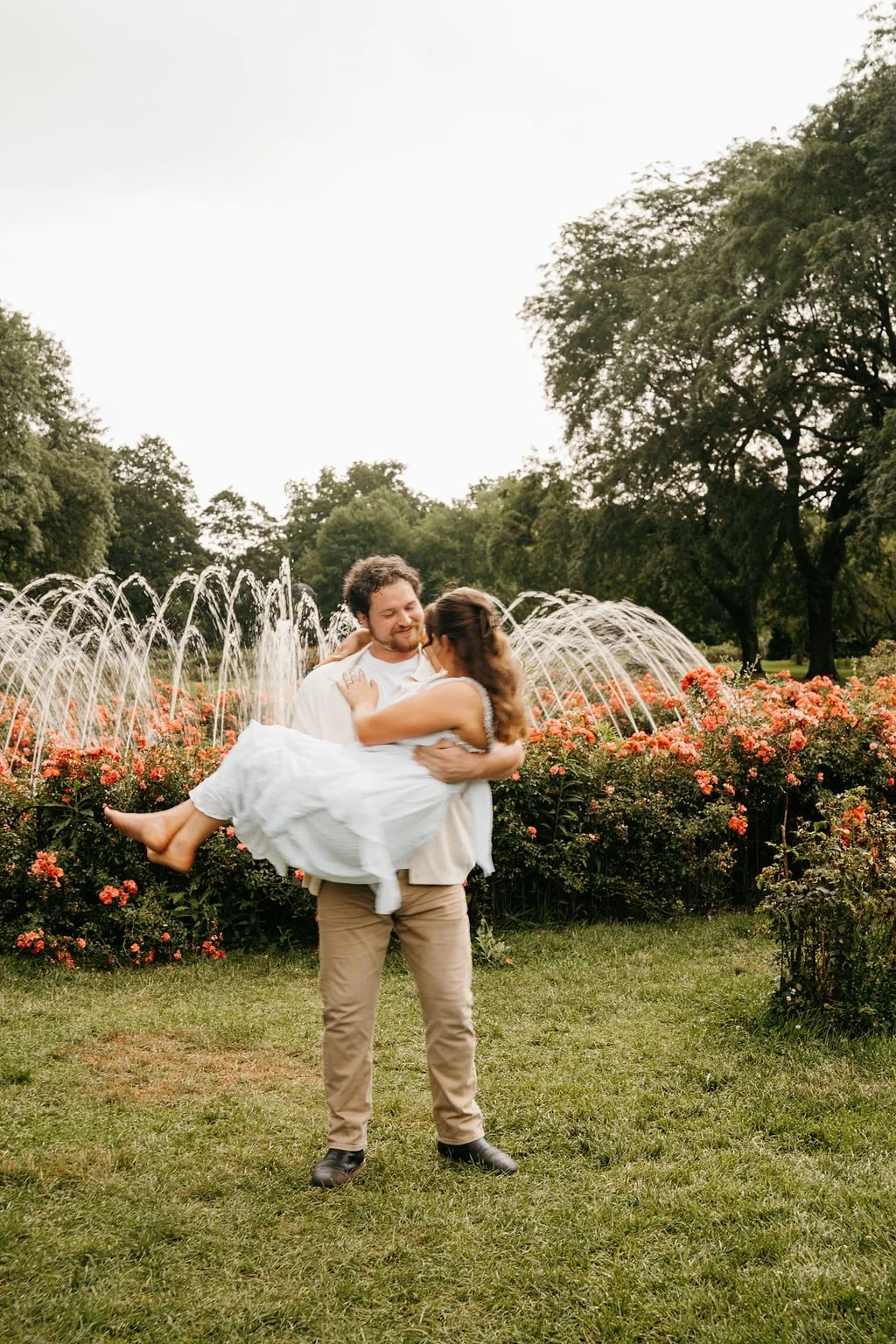 A man lifting a woman in a white dress in a garden with flowers and a fountain