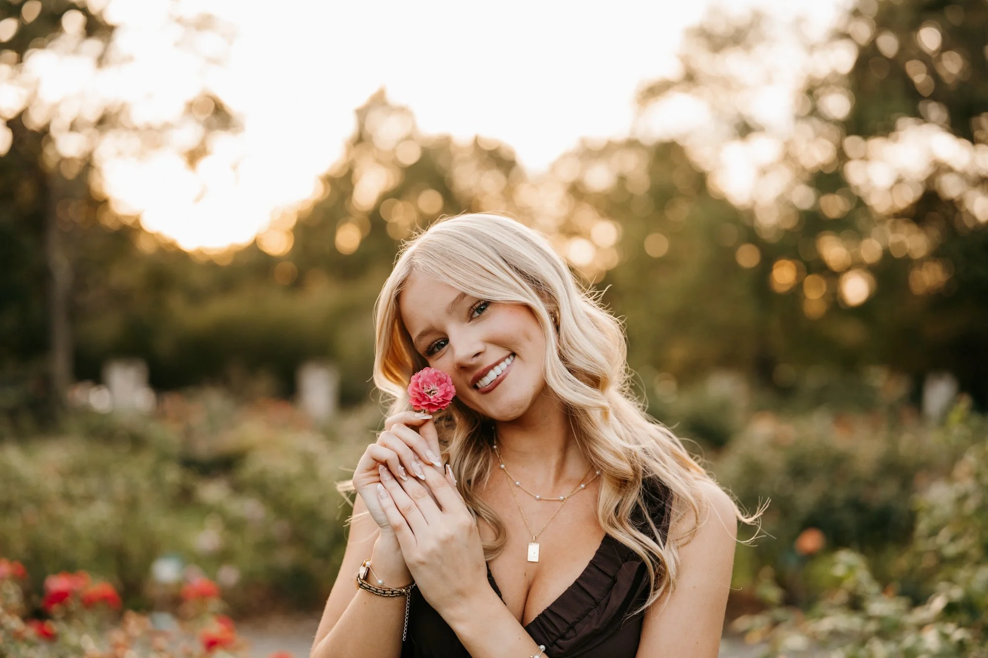 A woman with long blond hair smiling at the camera, holding a pink flower near her face outdoors during sunset.