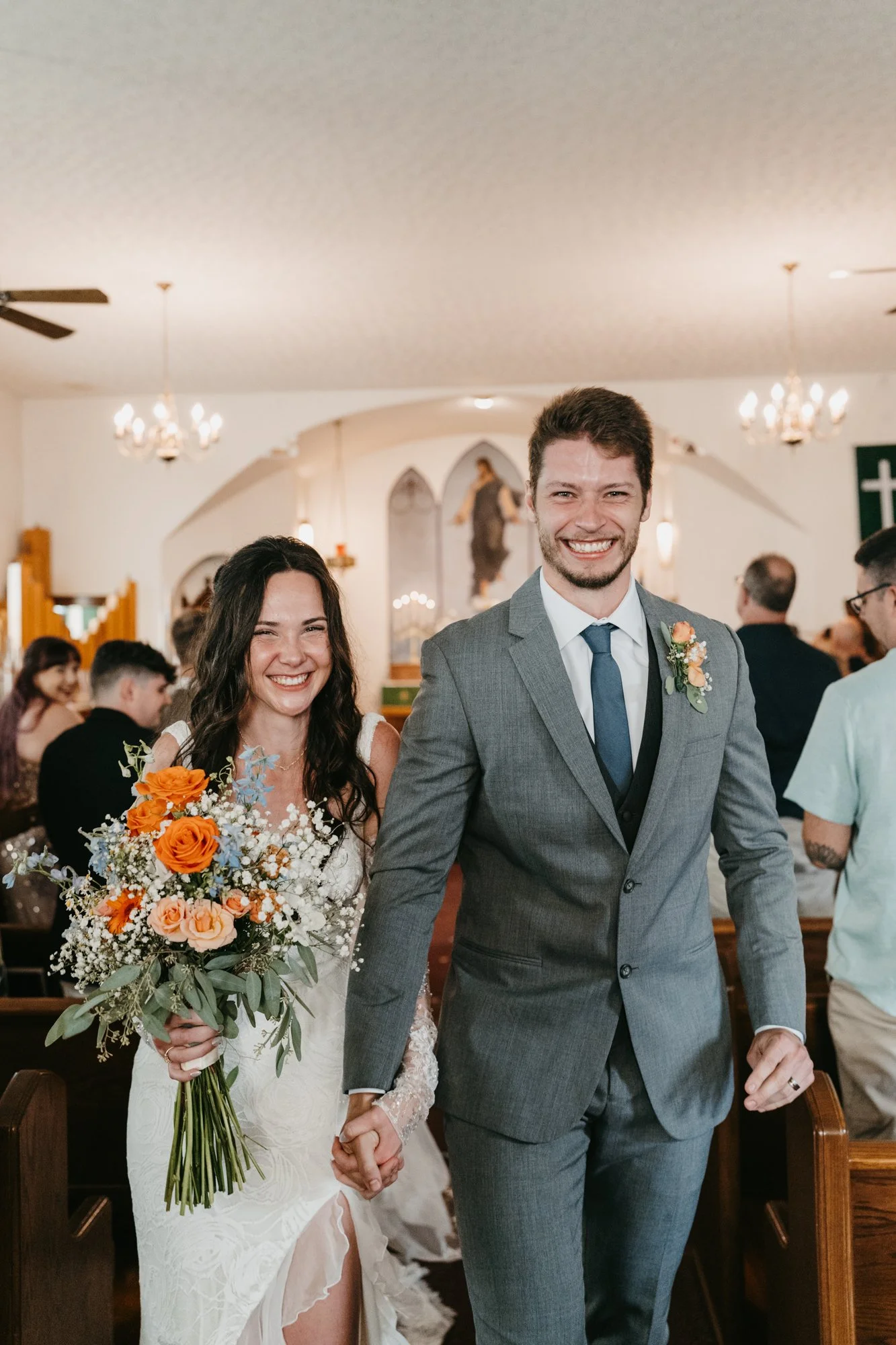 A newlywed couple walking down the aisle holding hands inside a church, smiling joyfully. The bride is holding a colorful bouquet of orange and peach roses, baby's breath, and greenery. The groom is dressed in a gray suit with a boutonniere on his la