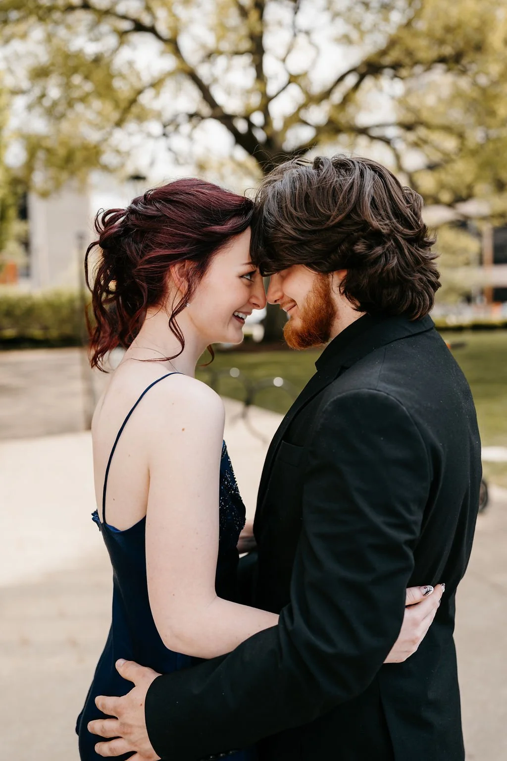 A couple is outside, face to face with foreheads touching, smiling, and holding each other. The woman has dark red hair and is wearing a dark blue dress, and the man has brown hair, a beard, and is wearing a black suit. They are standing in front of 