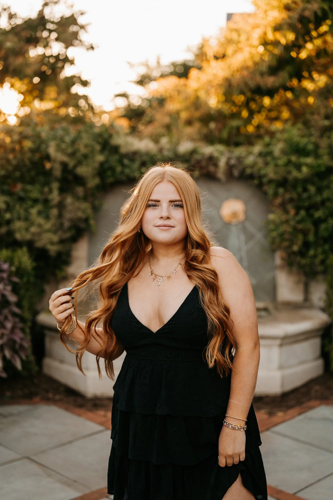 A woman with long, wavy red hair poses outdoors at sunset, wearing a black dress and layered gold jewelry, standing in front of a fountain and greenery.