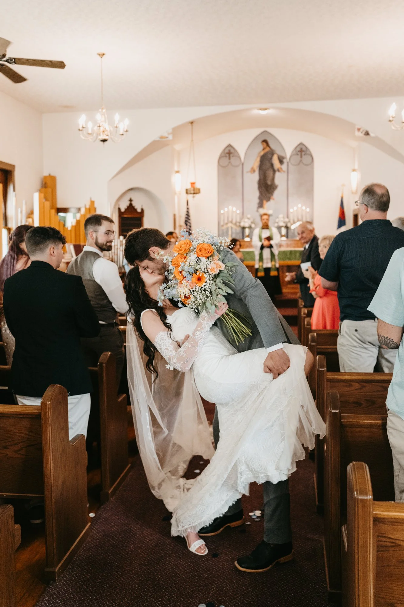 Bride and groom share a kiss during their wedding ceremony inside a church, with guests seated on pews and a religious altar in the background.