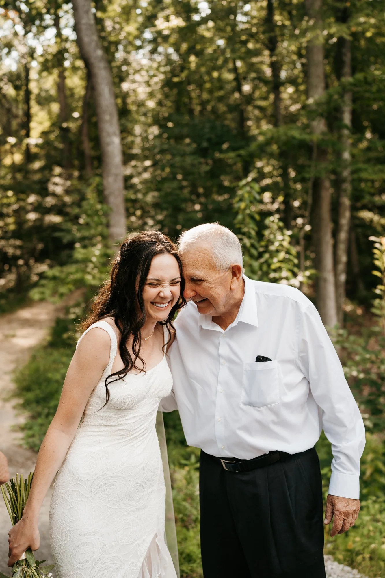 A young woman in a white wedding dress and an elderly man in a white shirt sharing a joyful moment outdoors in a wooded area.