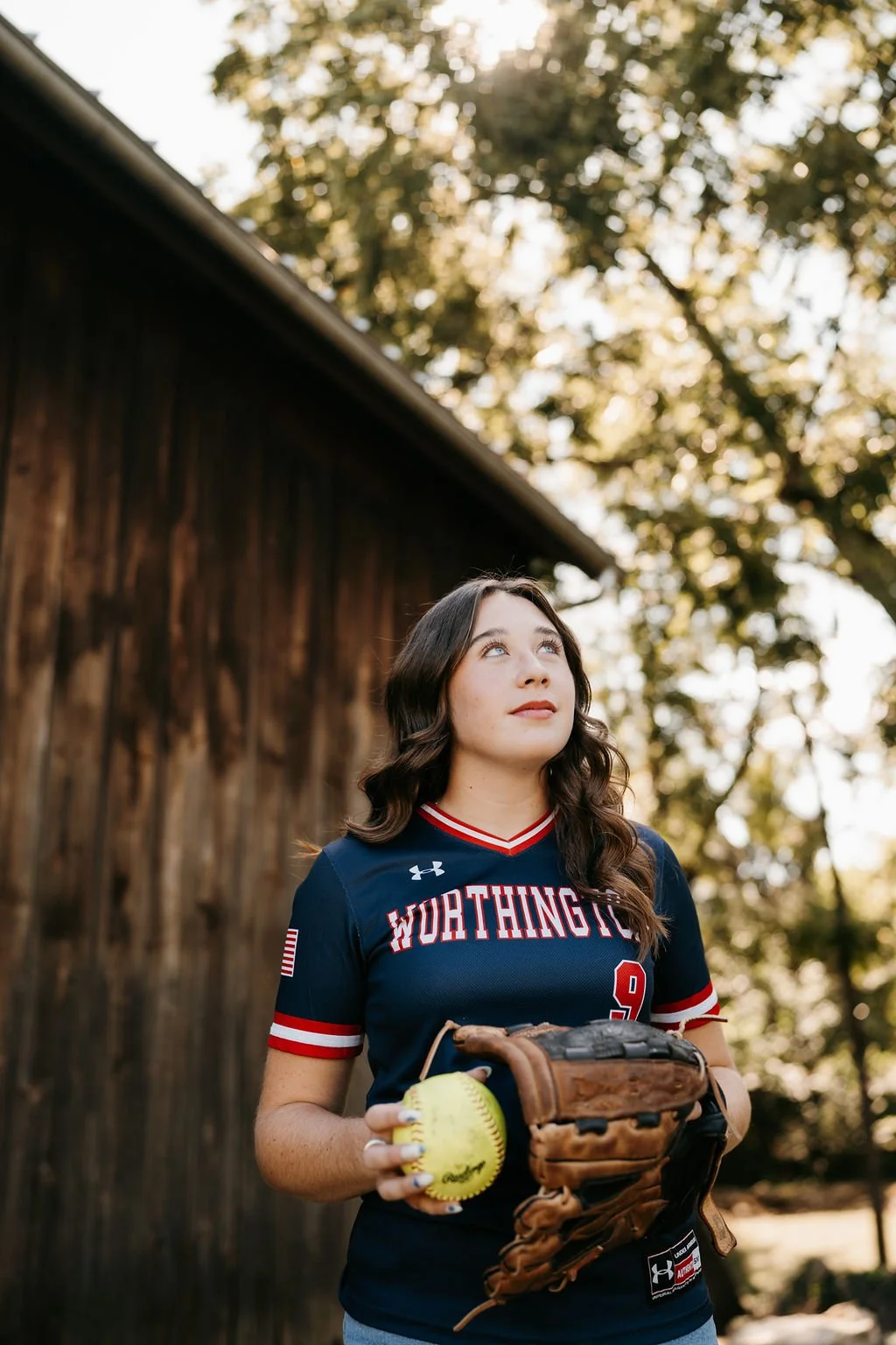 A young woman in a dark blue baseball jersey holding a yellow softball and a brown baseball glove outdoors during daytime, with trees in the background.