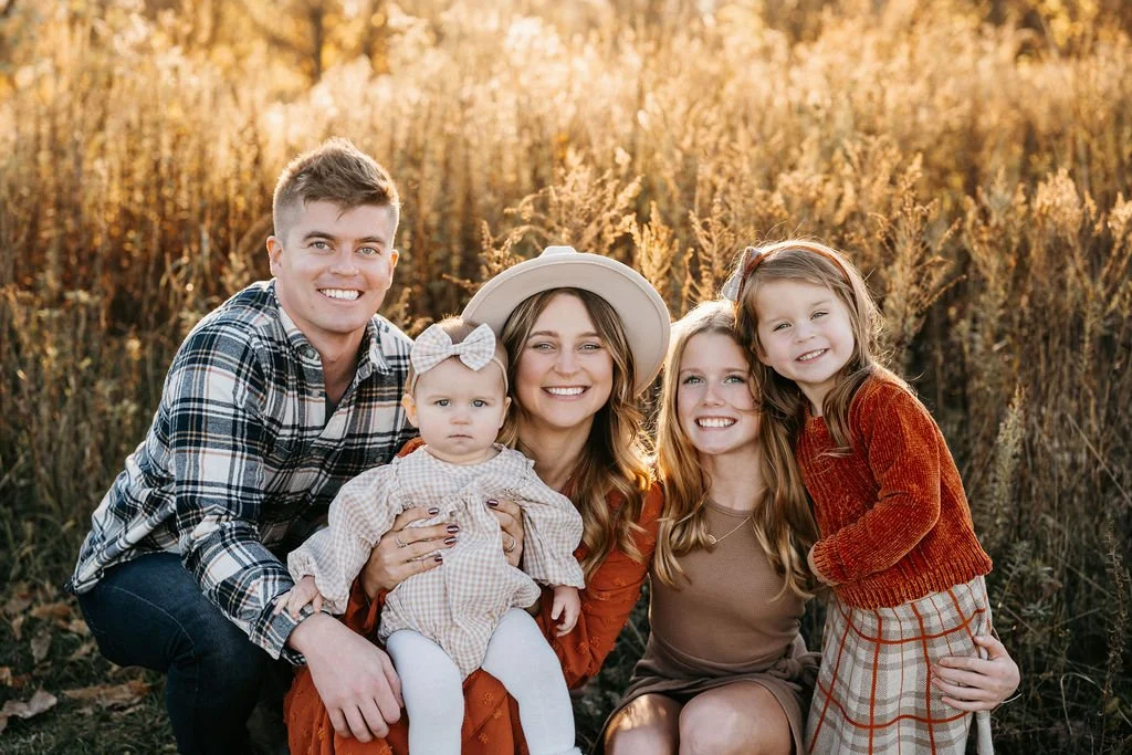 A family of five smiling in a field of tall grass during sunset, including a man, two women, and two young girls.